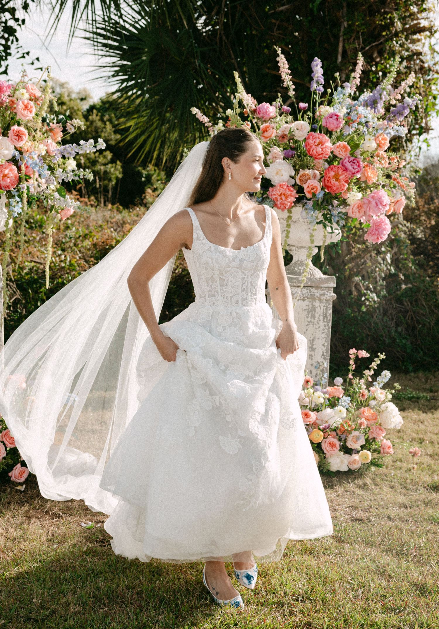 Bride in white lace tea-length wedding dress with veil, standing before colorful floral backdrop outdoors.