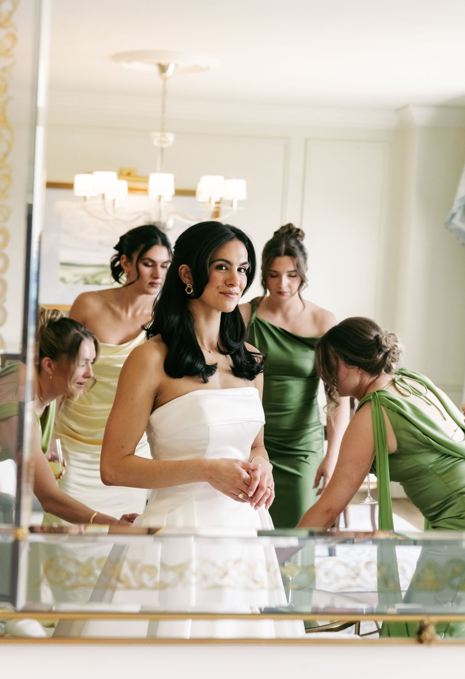 Bride in strapless white gown with bridesmaids in green and yellow dresses reflected in a large mirror.