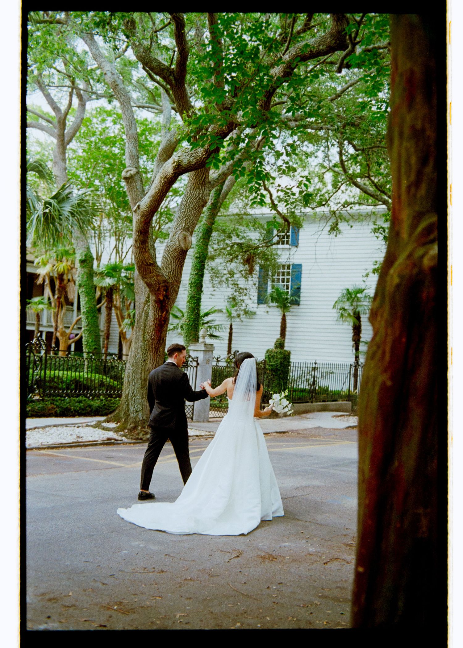 Bride and groom walking hand in hand on a tree-lined street, bride in flowing white gown with long train.