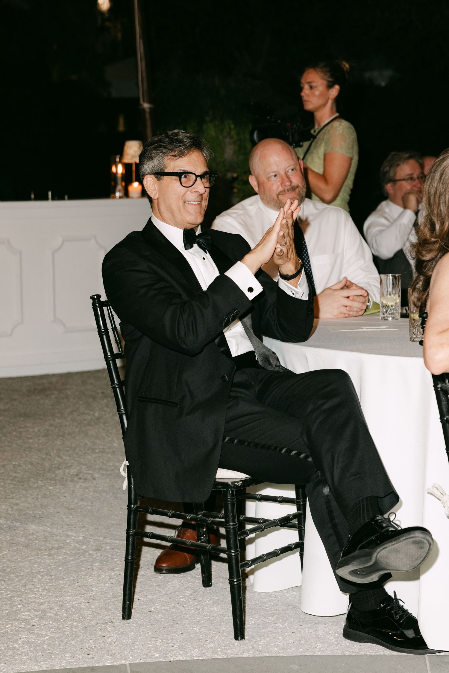 Smiling wedding guest in black tuxedo and glasses applauds during outdoor reception dinner.