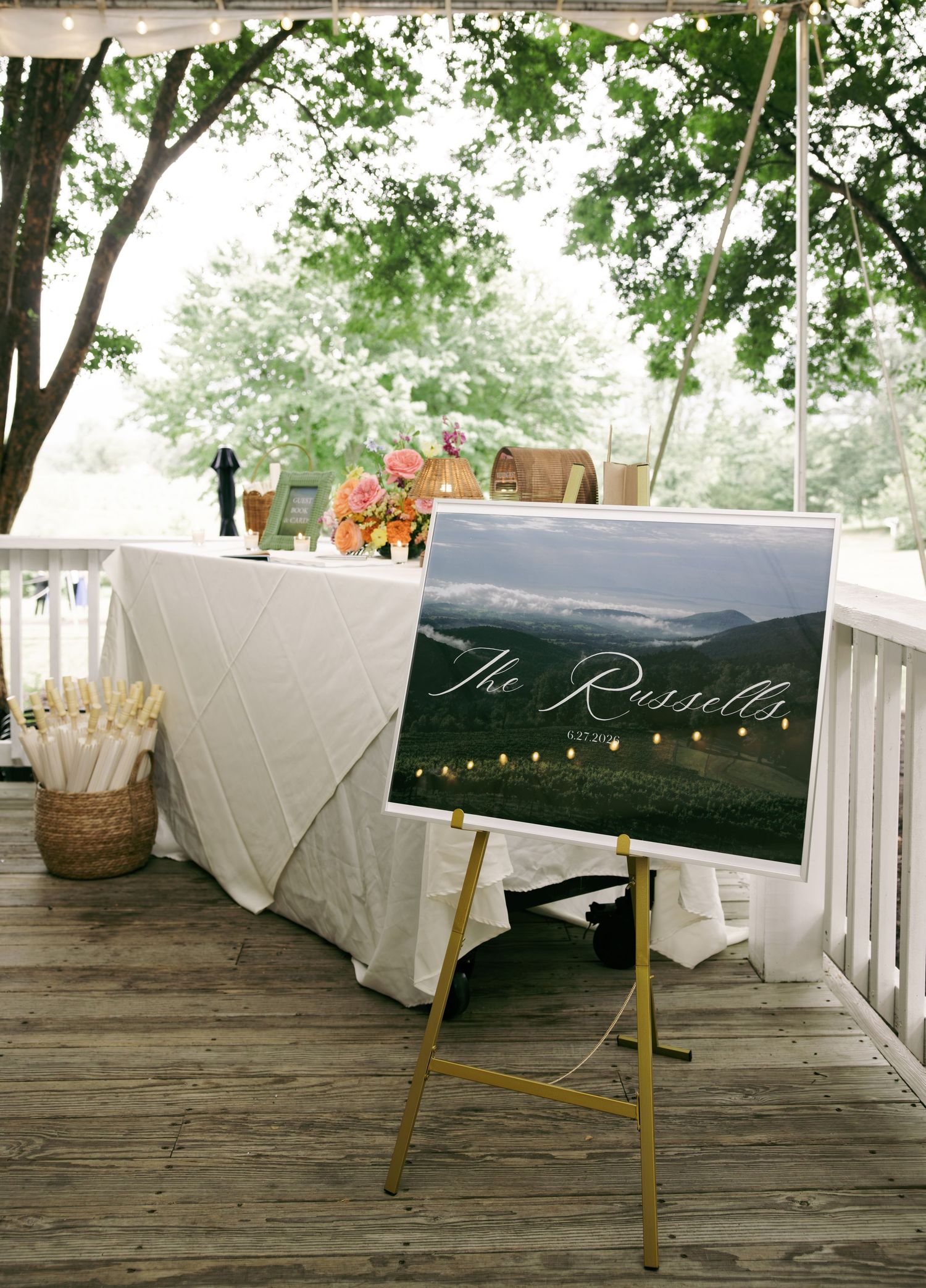 Wedding welcome sign reading The Russells on gold easel at decorated reception table on wooden deck.