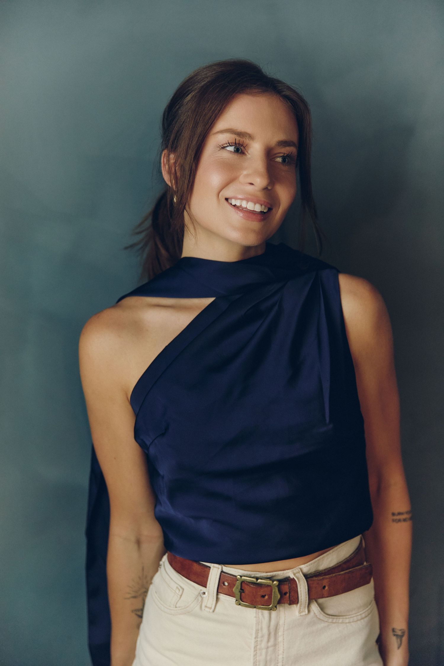 A person in a navy sleeveless top and white jeans poses against a dark background in a fashion photo series.