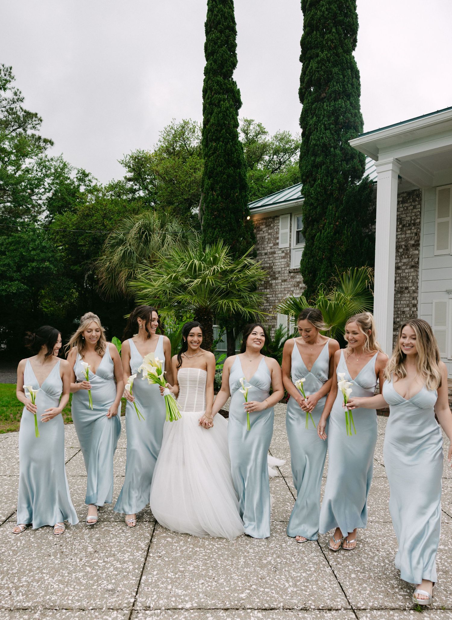 Bride in white gown walks with bridesmaids in light blue satin dresses holding white calla lilies outside a venue.