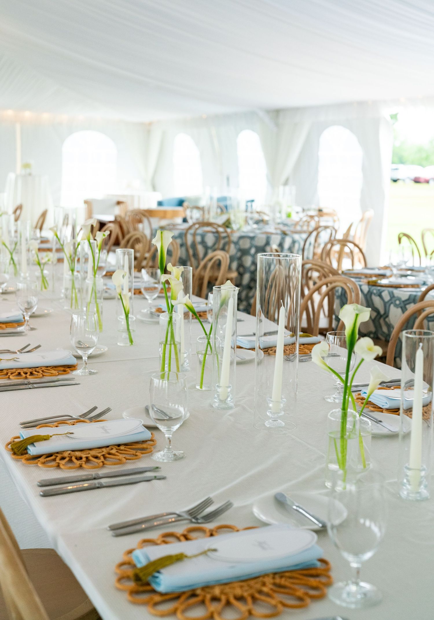 Elegant wedding reception table with white linens, wicker chargers, calla lily stems, and candles inside a white tent.