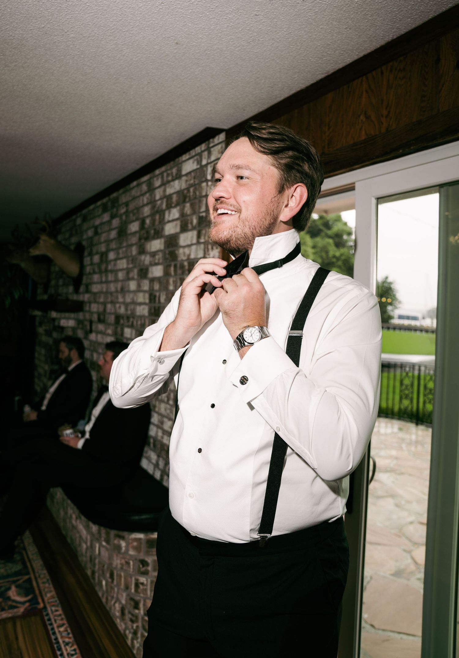 Groom in white tuxedo shirt and black suspenders adjusting his bow tie while getting ready for wedding.