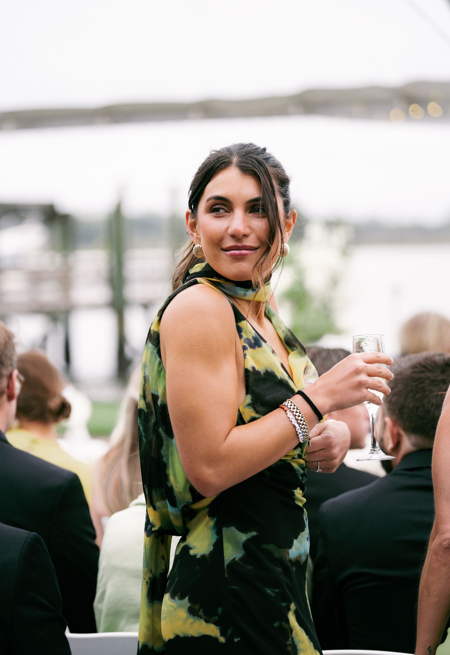 Female wedding guest in green floral dress holding champagne glass at waterfront outdoor reception.
