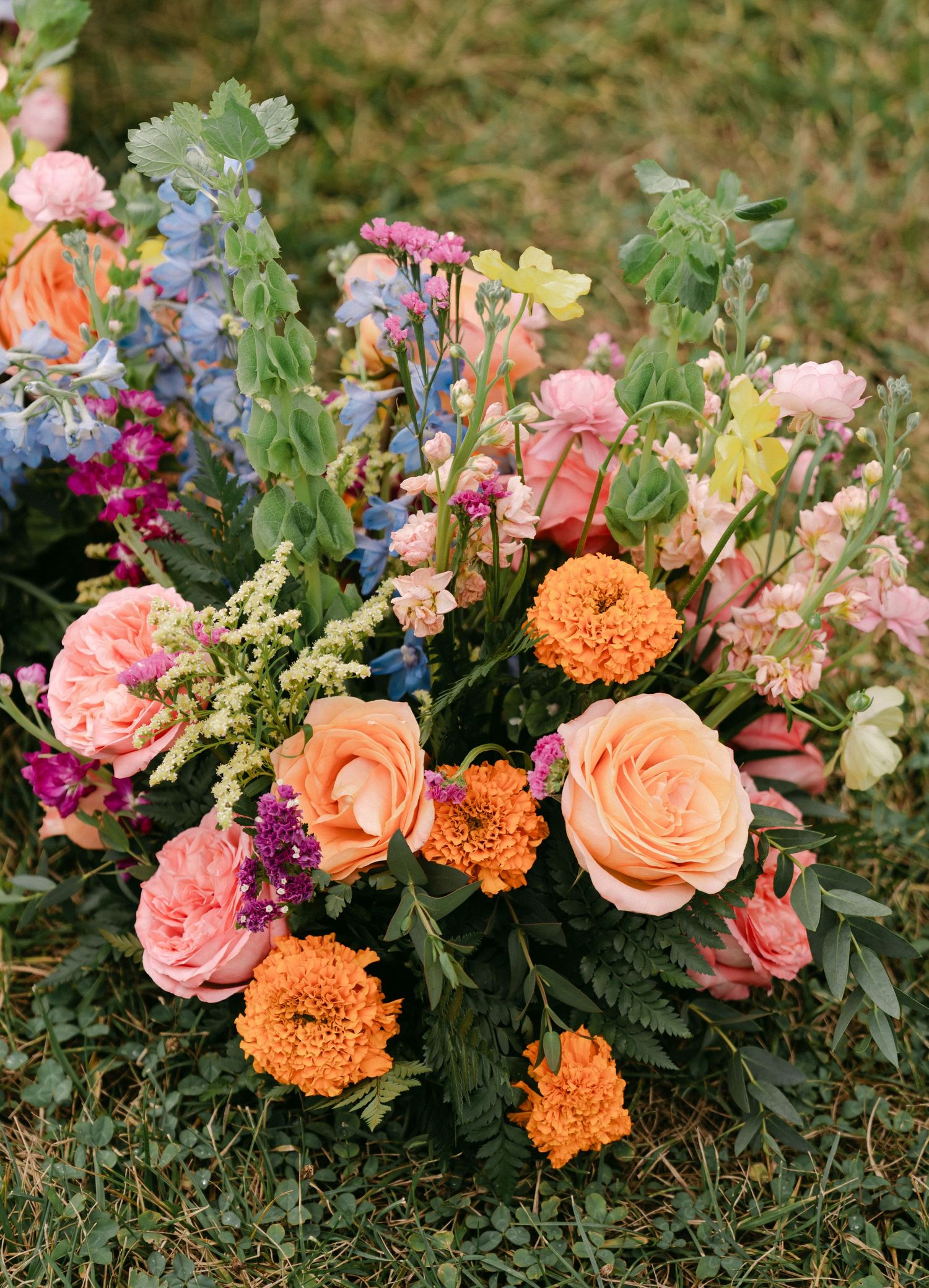 Vibrant mixed floral arrangement with orange marigolds, peach roses, pink blooms, and lush greenery on grass.