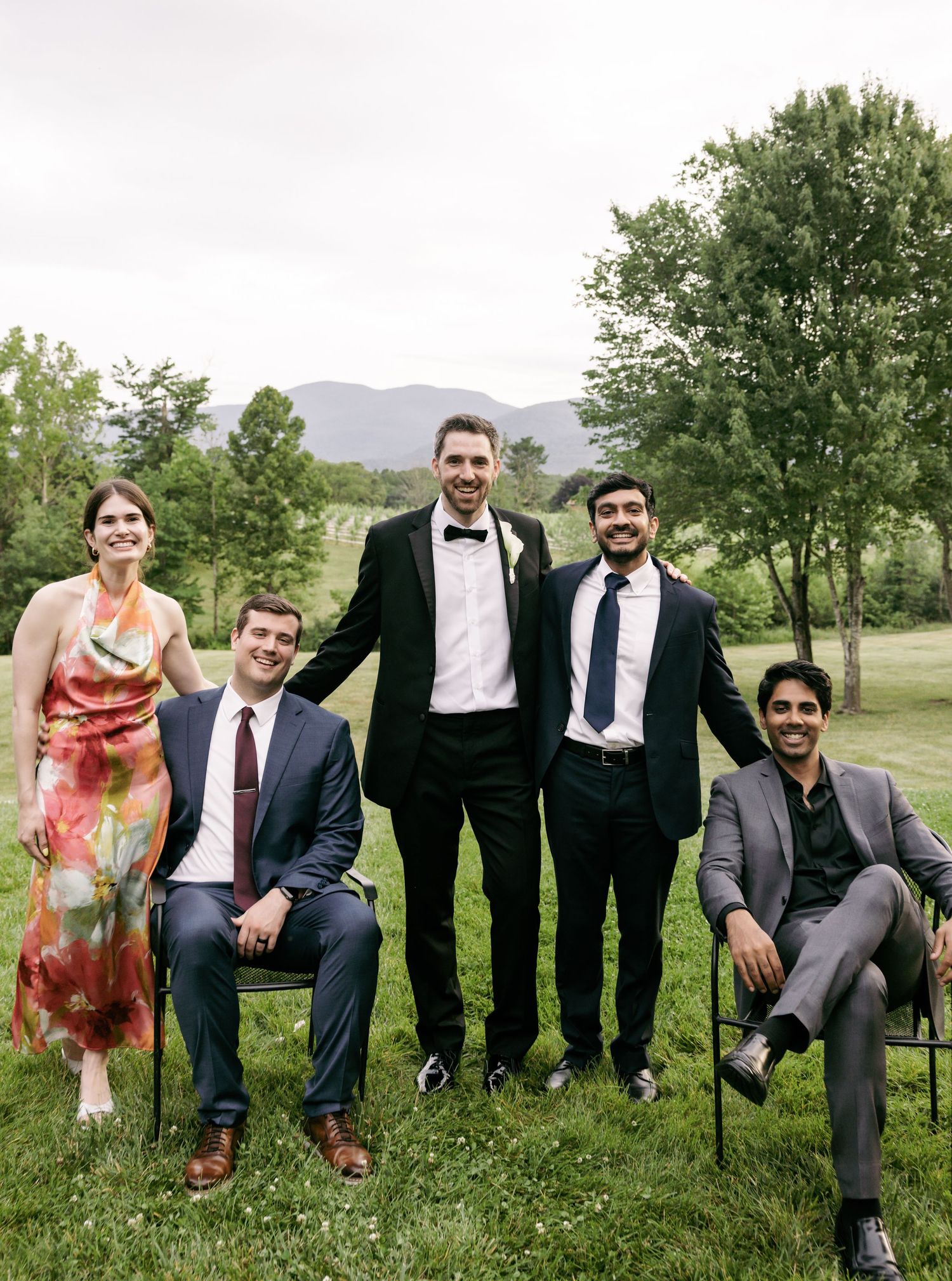 Groom with wedding guests posing outdoors on green lawn with mountain views in background.