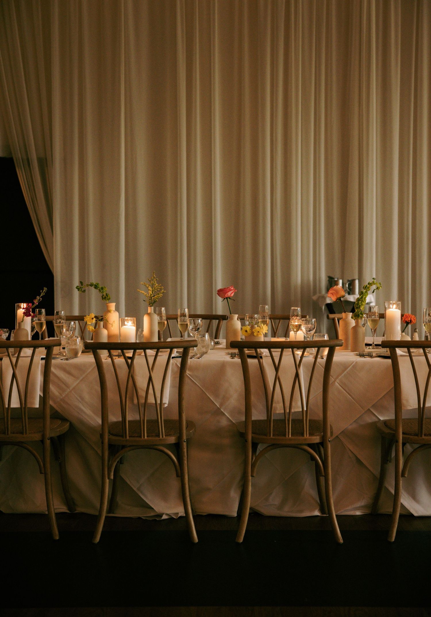 Elegant wedding reception table with gold chairs, candles, and floral centerpieces against draped white curtains.