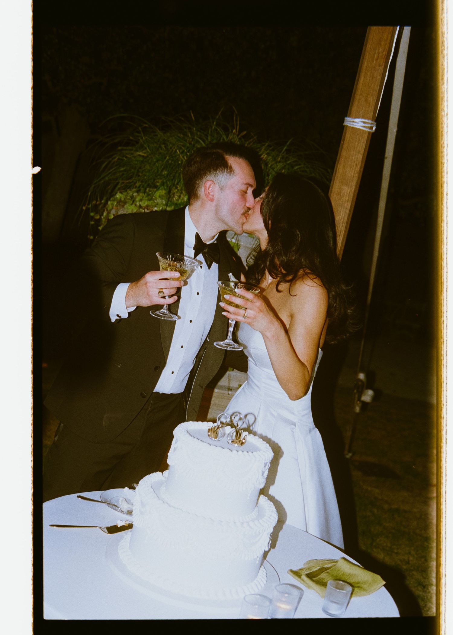 Newlyweds kissing while holding champagne glasses beside a white tiered wedding cake.