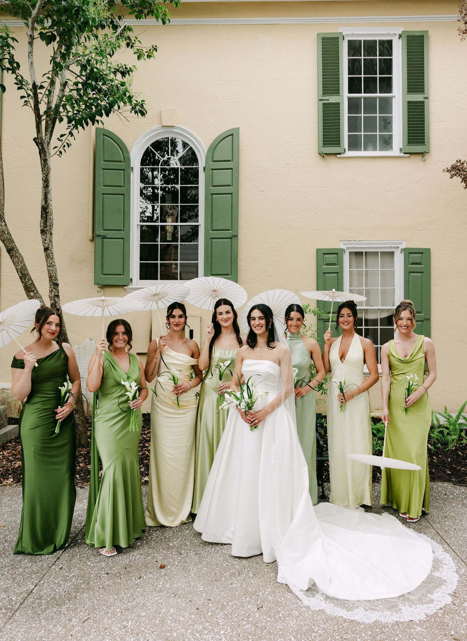 Bride in white gown poses with bridesmaids in green satin dresses holding parasols outside a yellow building.