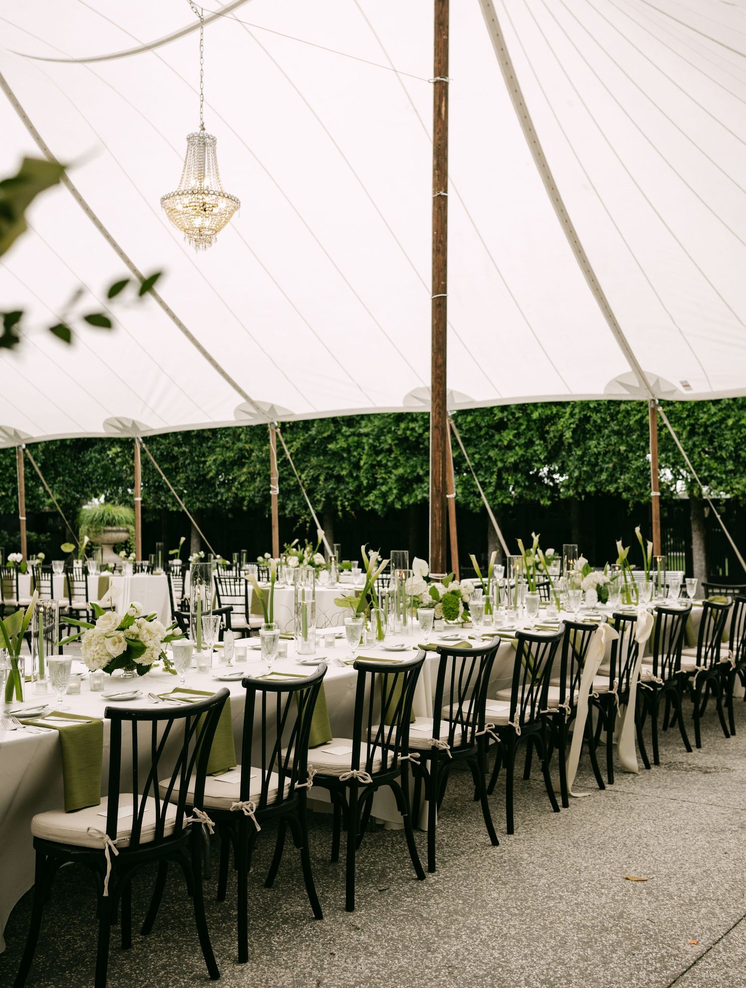 Long wedding reception tables set under a white sailcloth tent with black chairs, crystal chandelier, and green centerpieces.