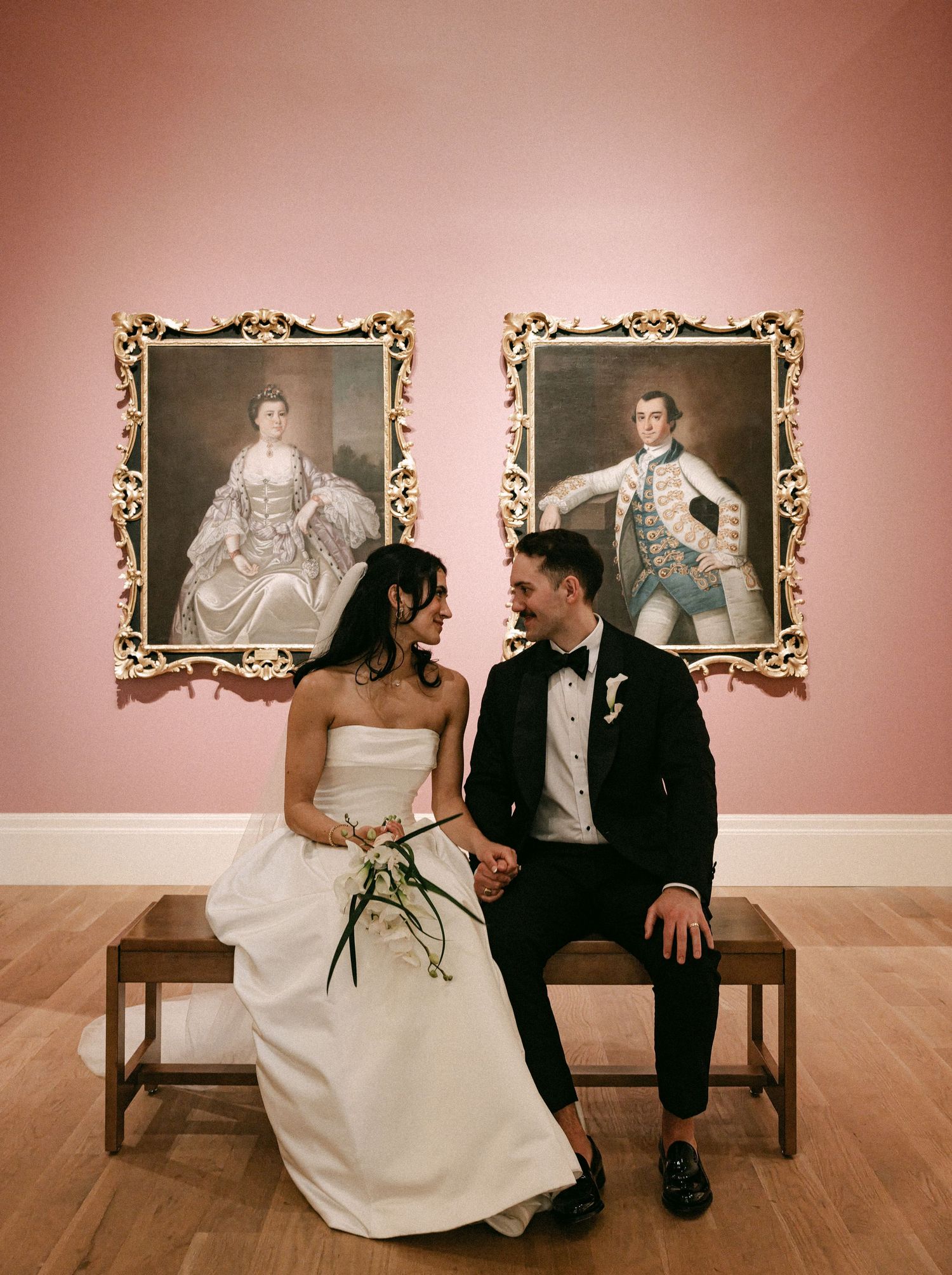 Bride and groom sit together in art museum in front of ornate gold-framed classical portrait paintings.