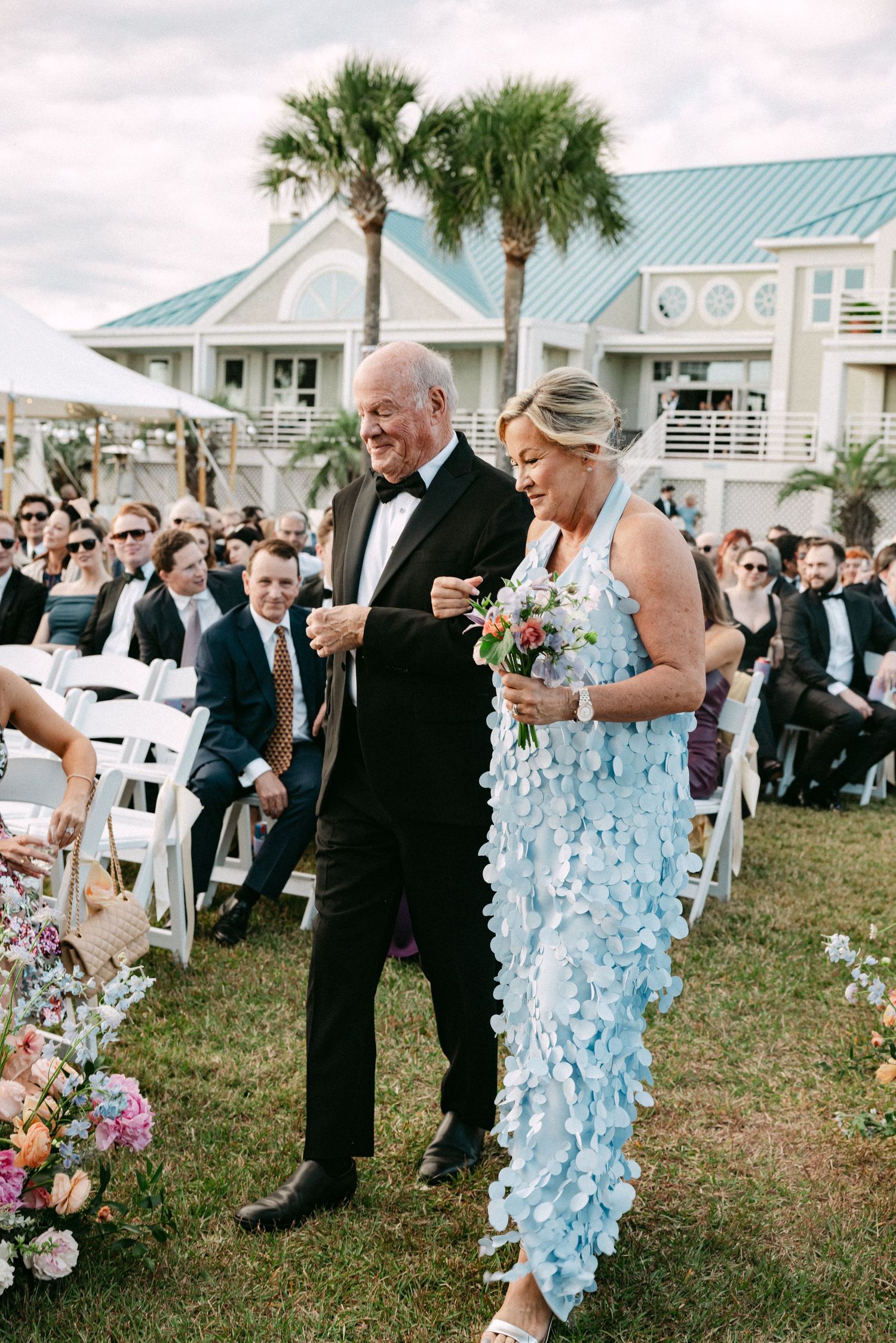 Couple walking down wedding aisle, woman in blue petal dress holding bouquet outdoors.