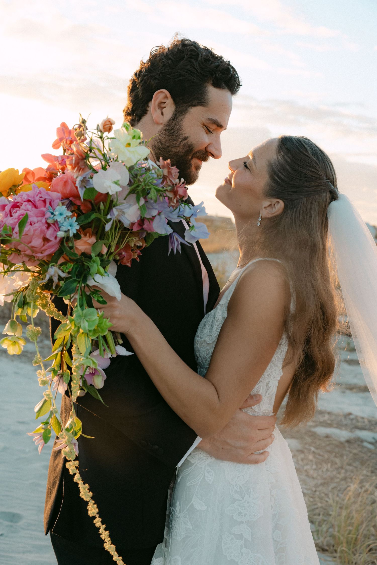 Bride and groom embrace romantically holding colorful cascading bouquet at sunset during outdoor wedding photo session.