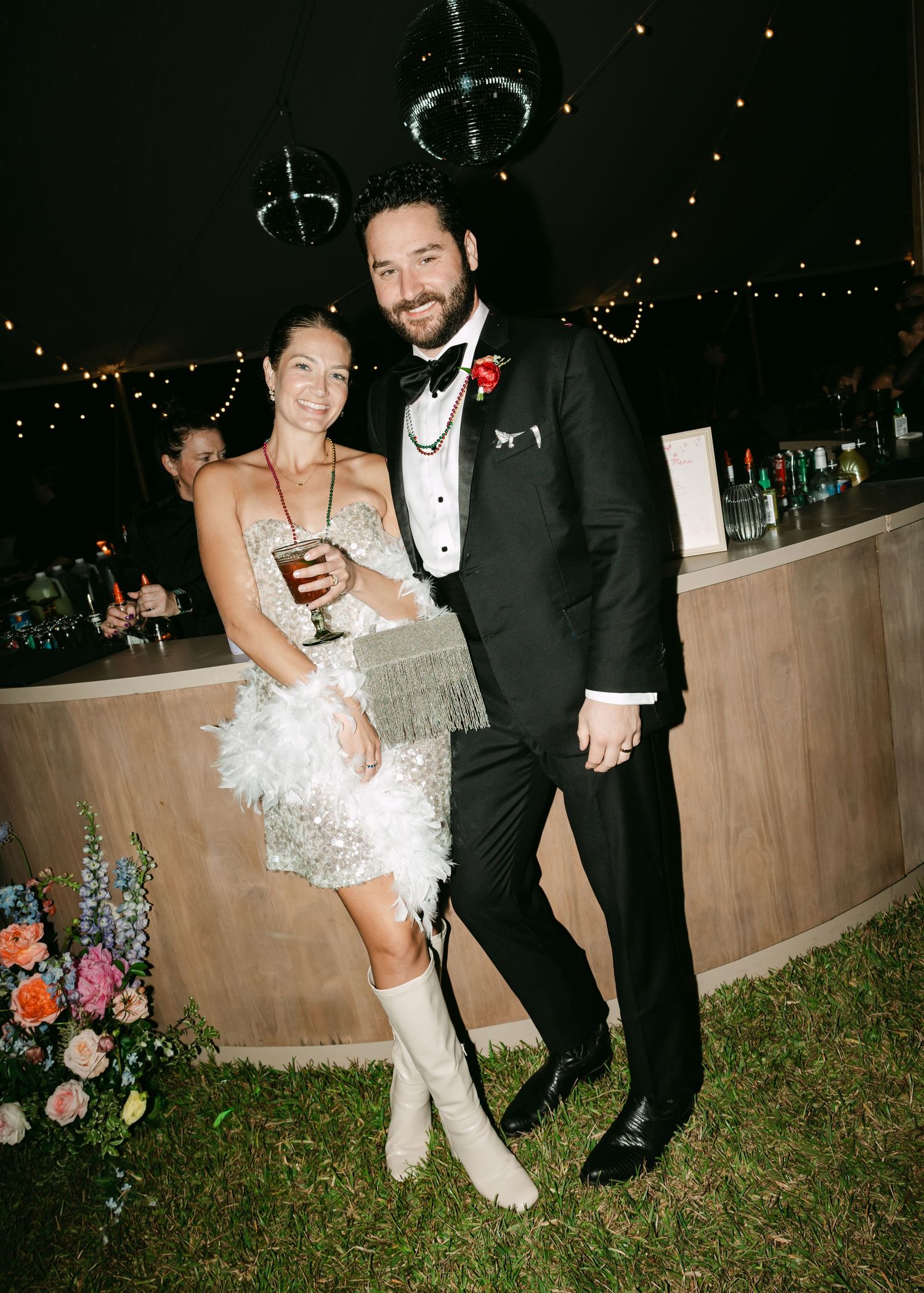 Bride in sequin fringe dress and groom in black tuxedo posing at outdoor wedding reception.