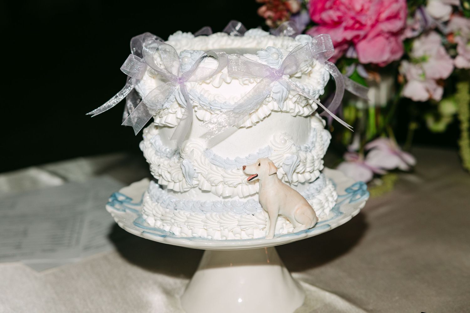Small white wedding cake with lace details and animal figurine displayed on white cake stand.