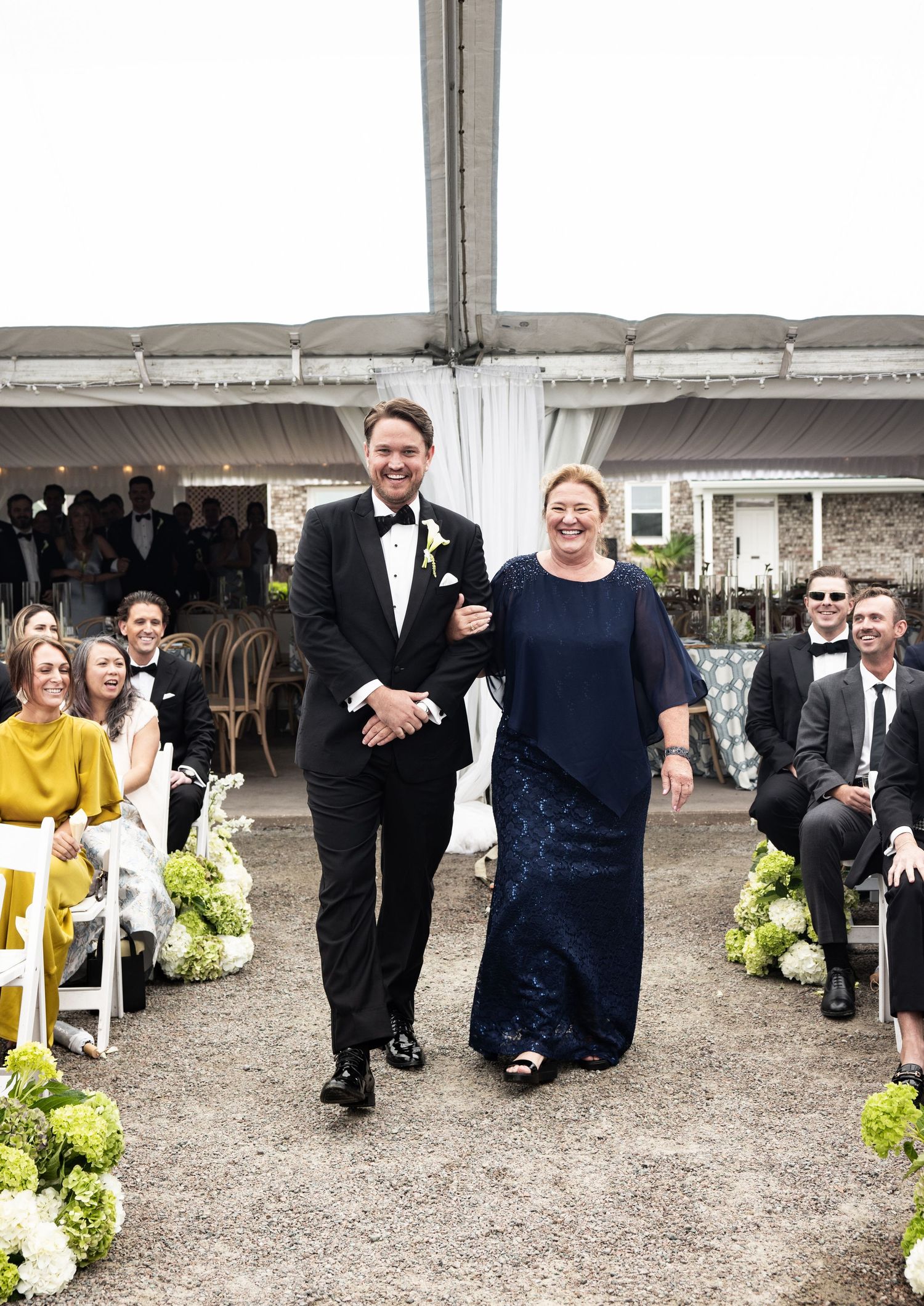 Groom walks down wedding aisle with mother in navy lace dress under white tent with seated guests.