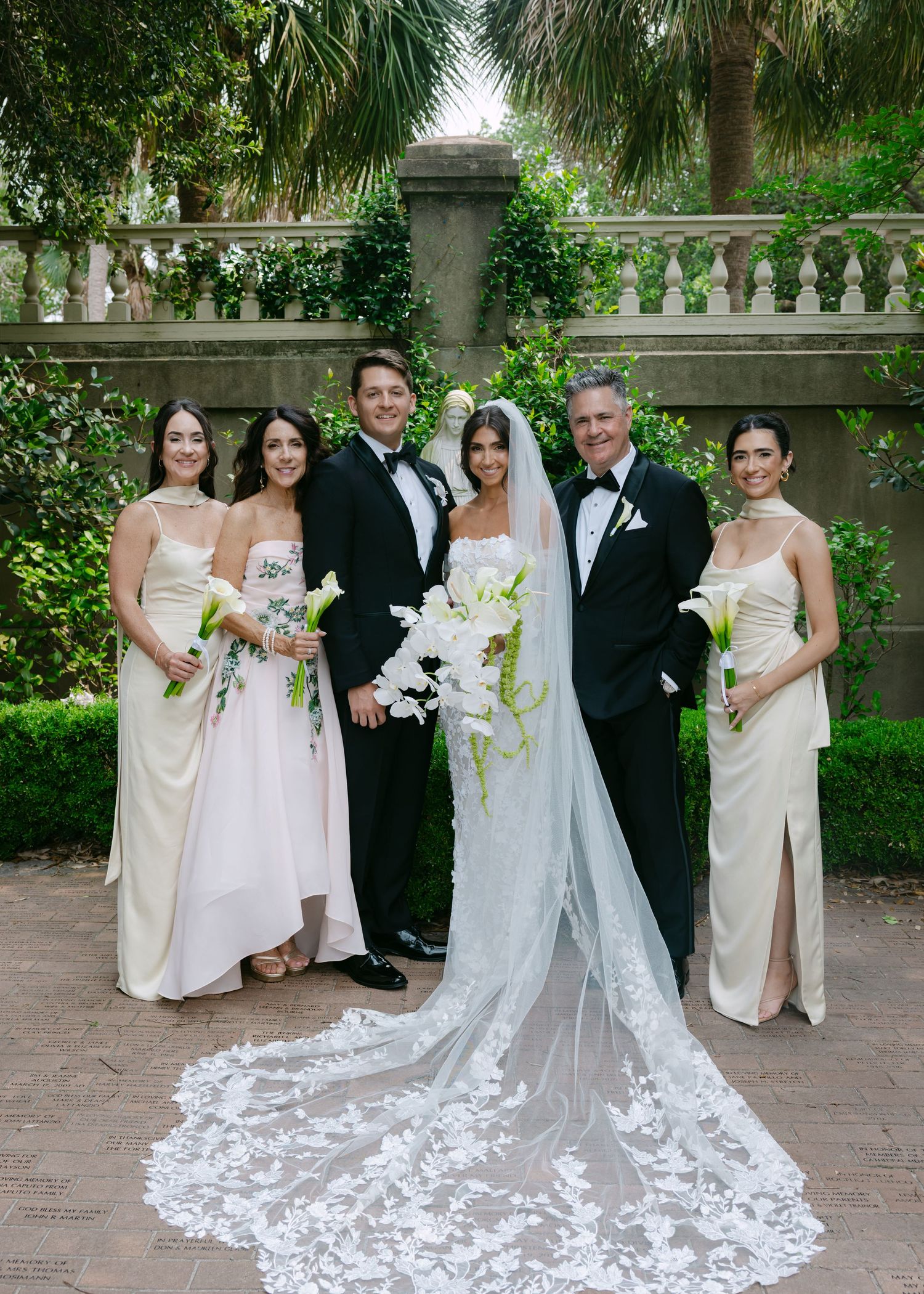 Wedding party in cream dresses and black tuxedos pose together on brick pathway surrounded by palm trees.