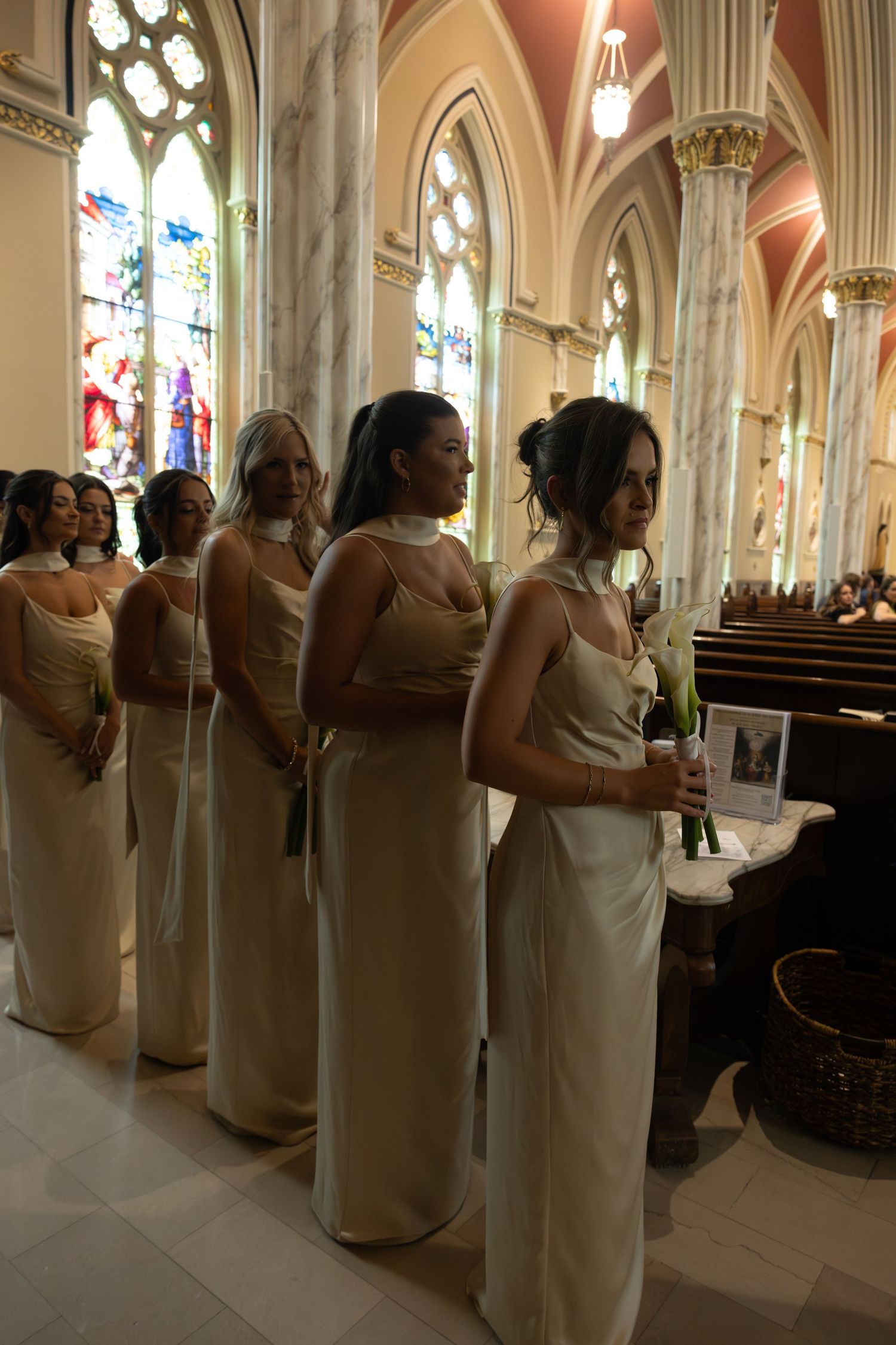 Bridesmaids in cream-colored dresses stand in line inside a cathedral with stained glass windows.