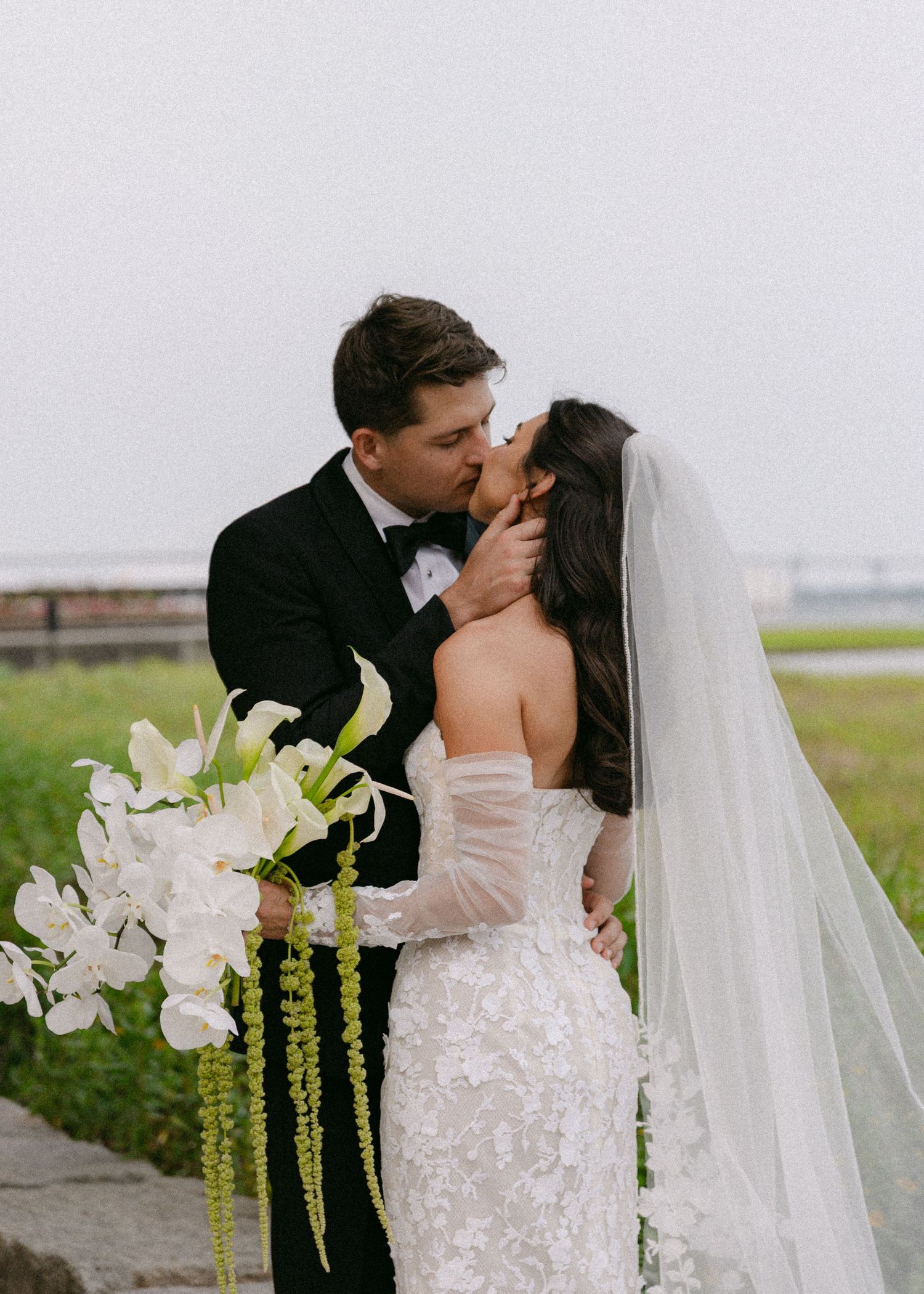 Romantic wedding portrait with white orchid bouquet and flowing veil against natural backdrop.