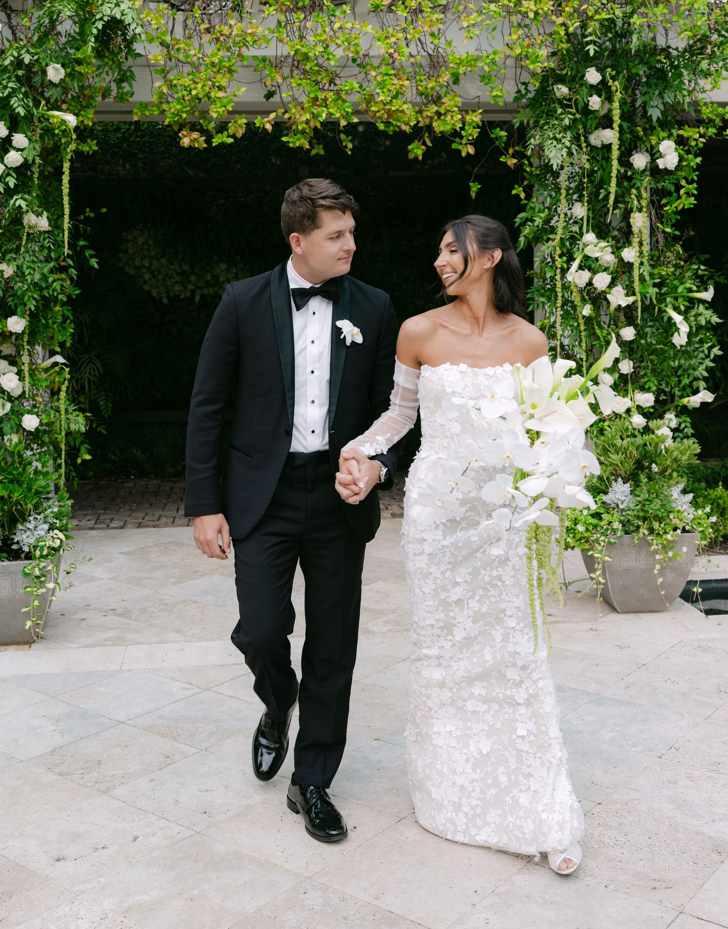 Newlyweds walk together on a stone pathway with elegant white floral arrangements and greenery overhead.