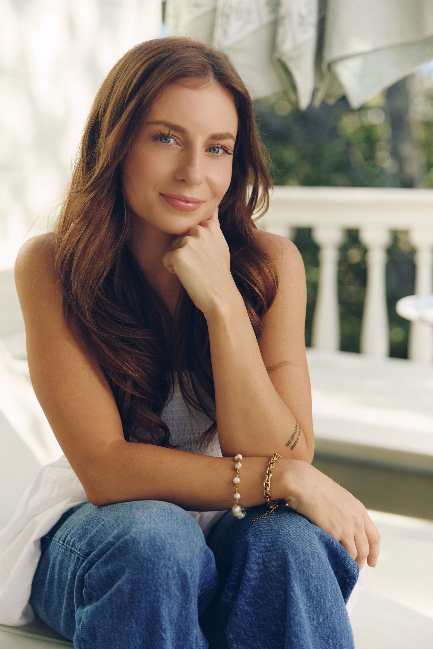 Woman in casual attire sitting on white porch with contemplative pose in natural lighting.