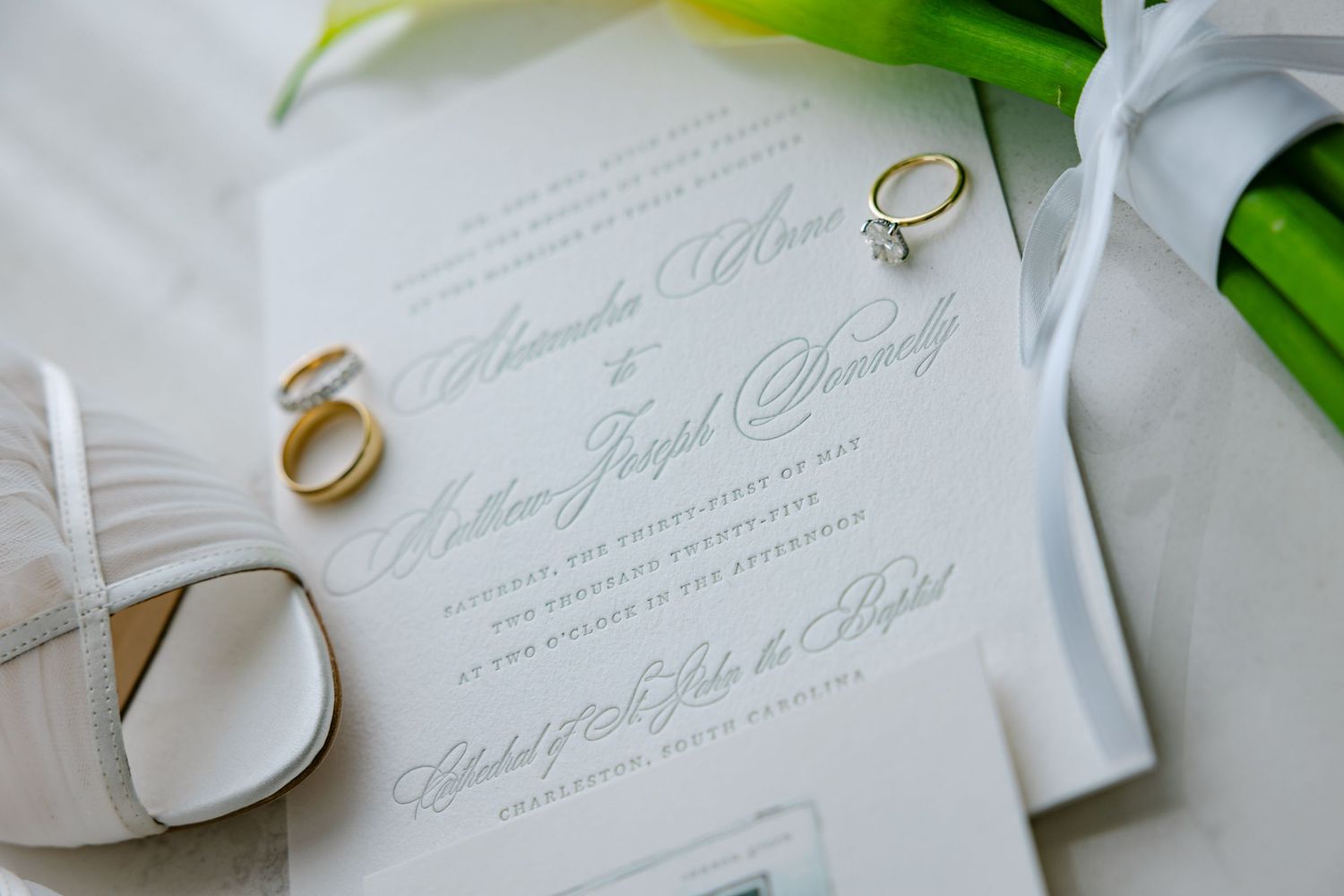 Elegant wedding stationery with gold rings and calla lily bouquet on white surface.