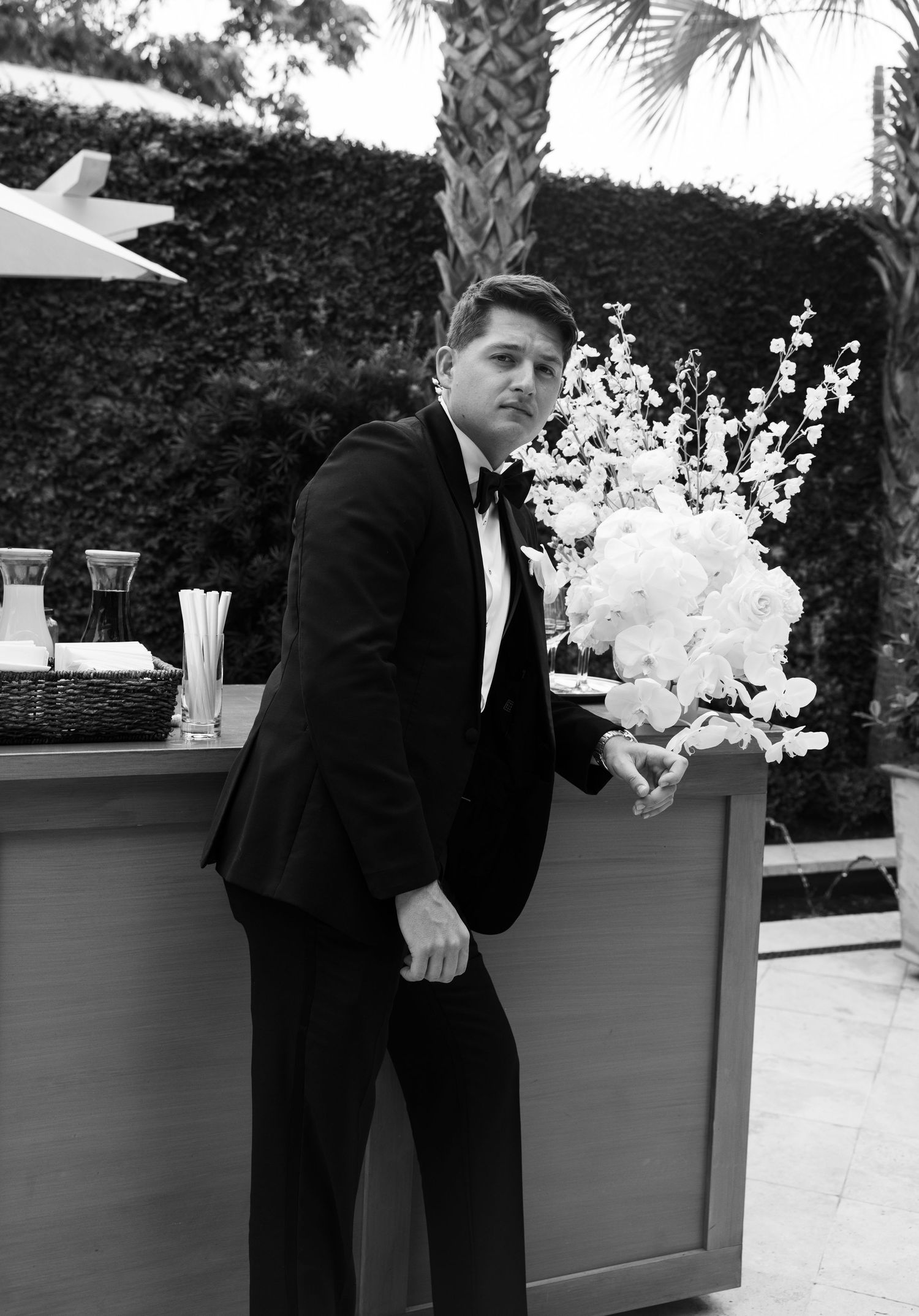 Groom in black tuxedo strikes a dramatic pose against a landscaped garden backdrop.