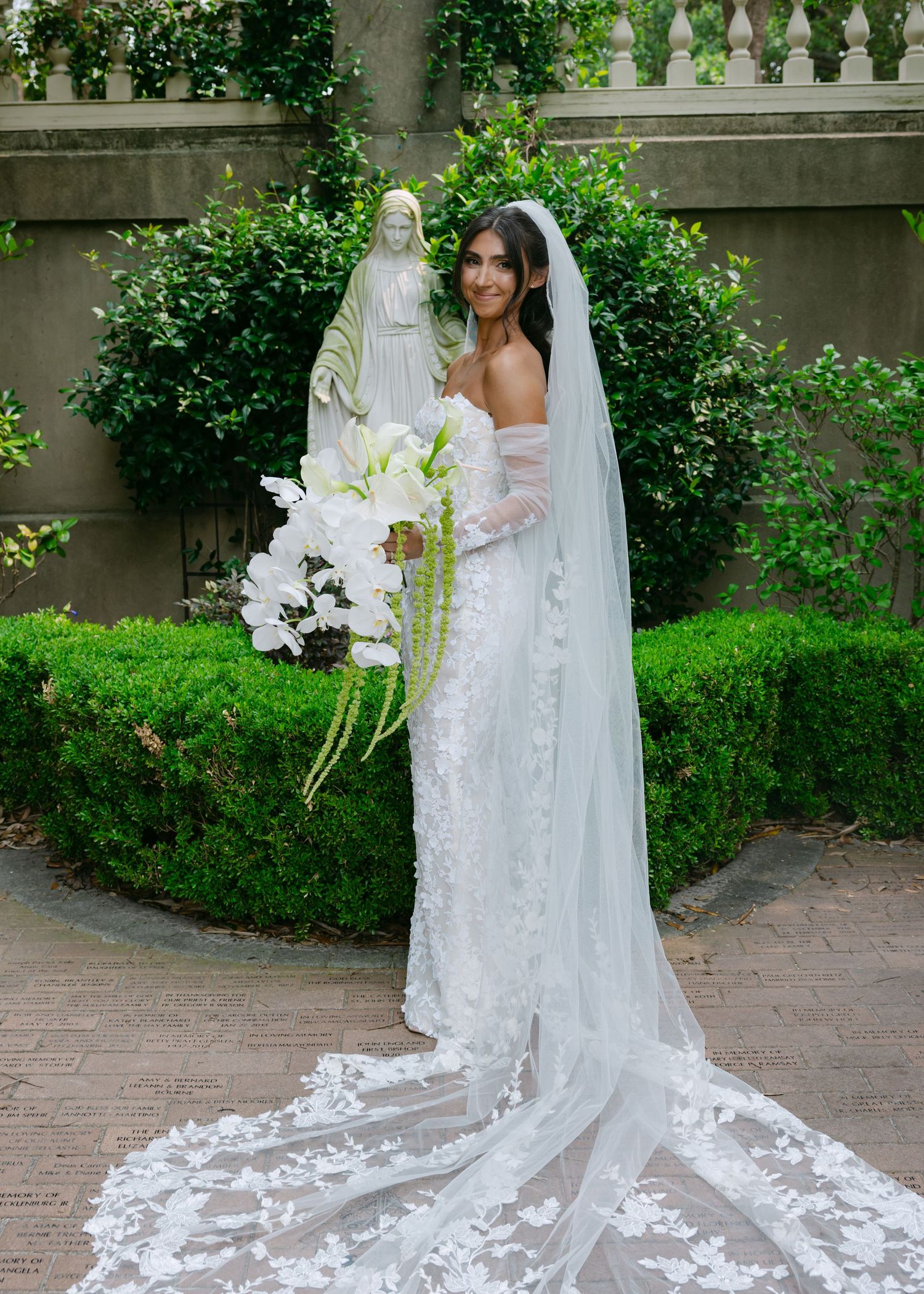Detailed view of an elegant white lace wedding dress train on brick pavers surrounded by greenery.