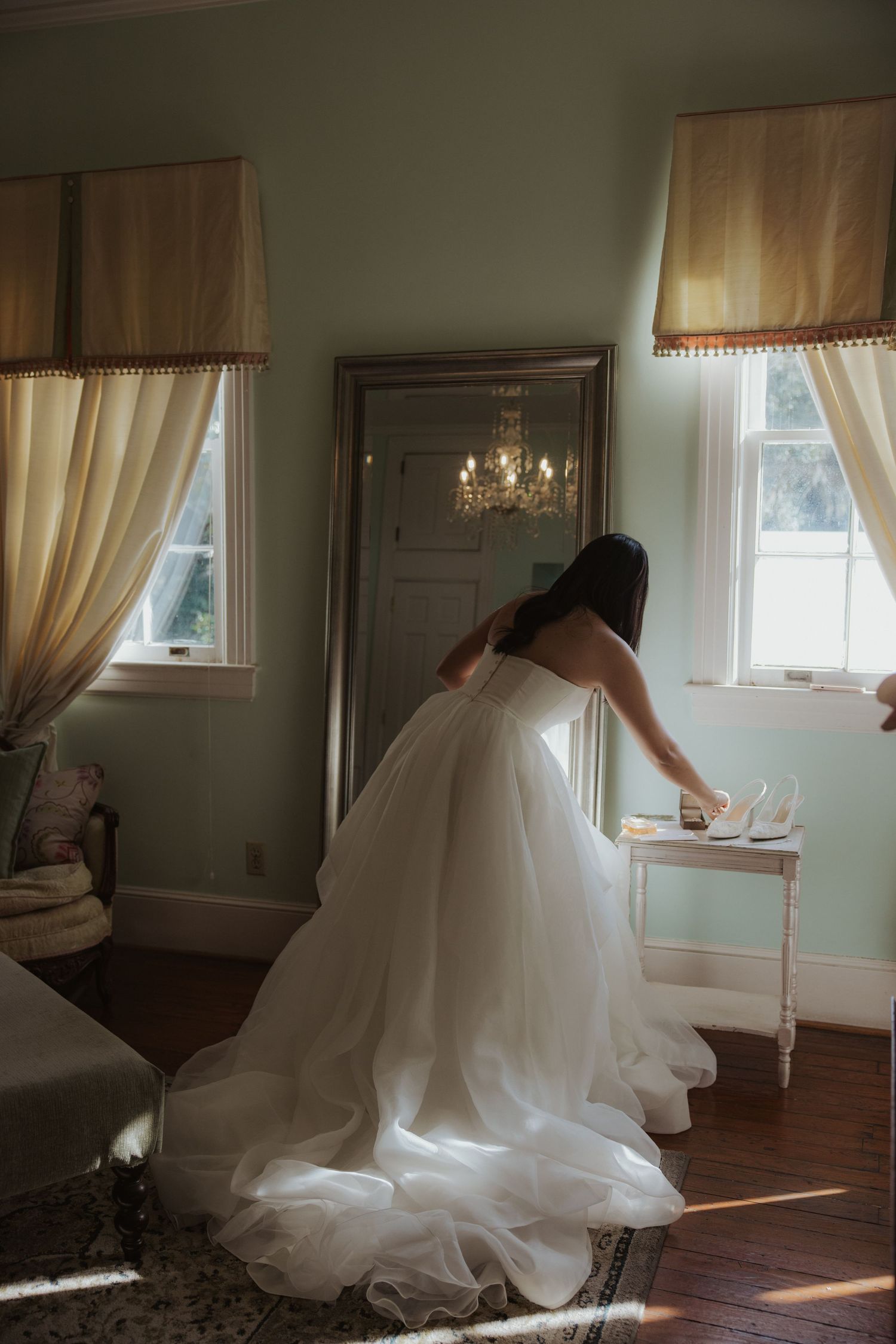 Flowing white wedding gown creates dramatic silhouette in sunlit room with cream curtains.