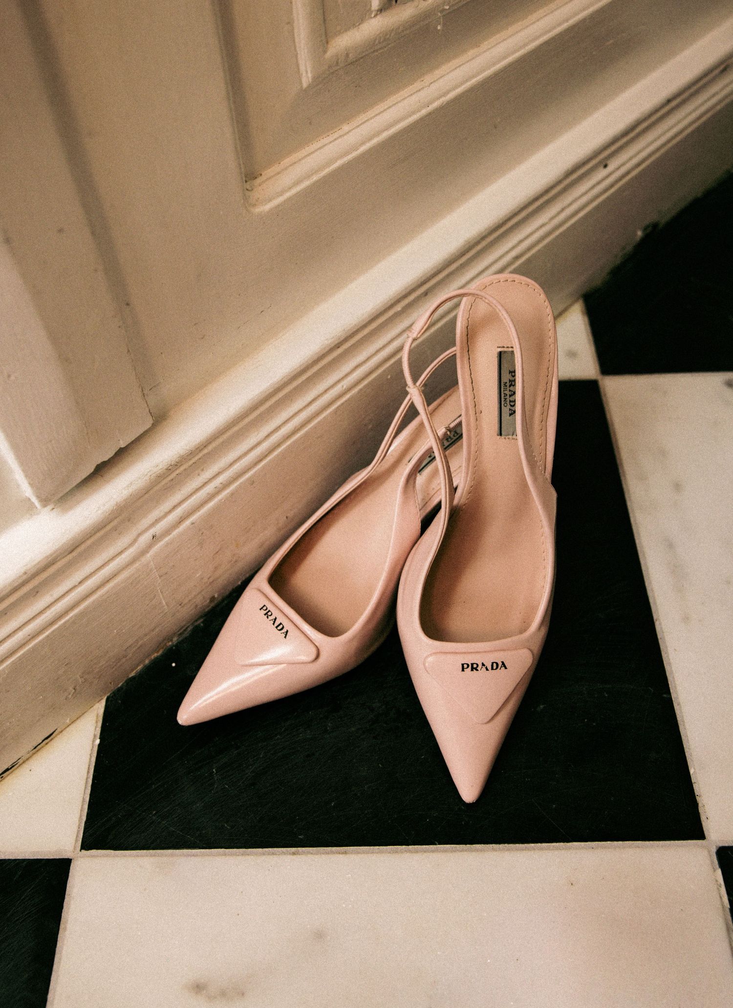 Prada blush pink patent leather pointed-toe slingback heels placed on a black and white checkered floor.