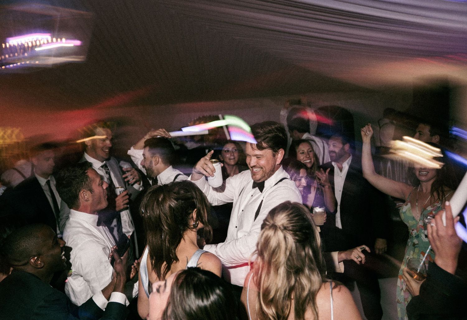 Guests dancing and celebrating at a lively wedding reception party under a tent with colorful lights.