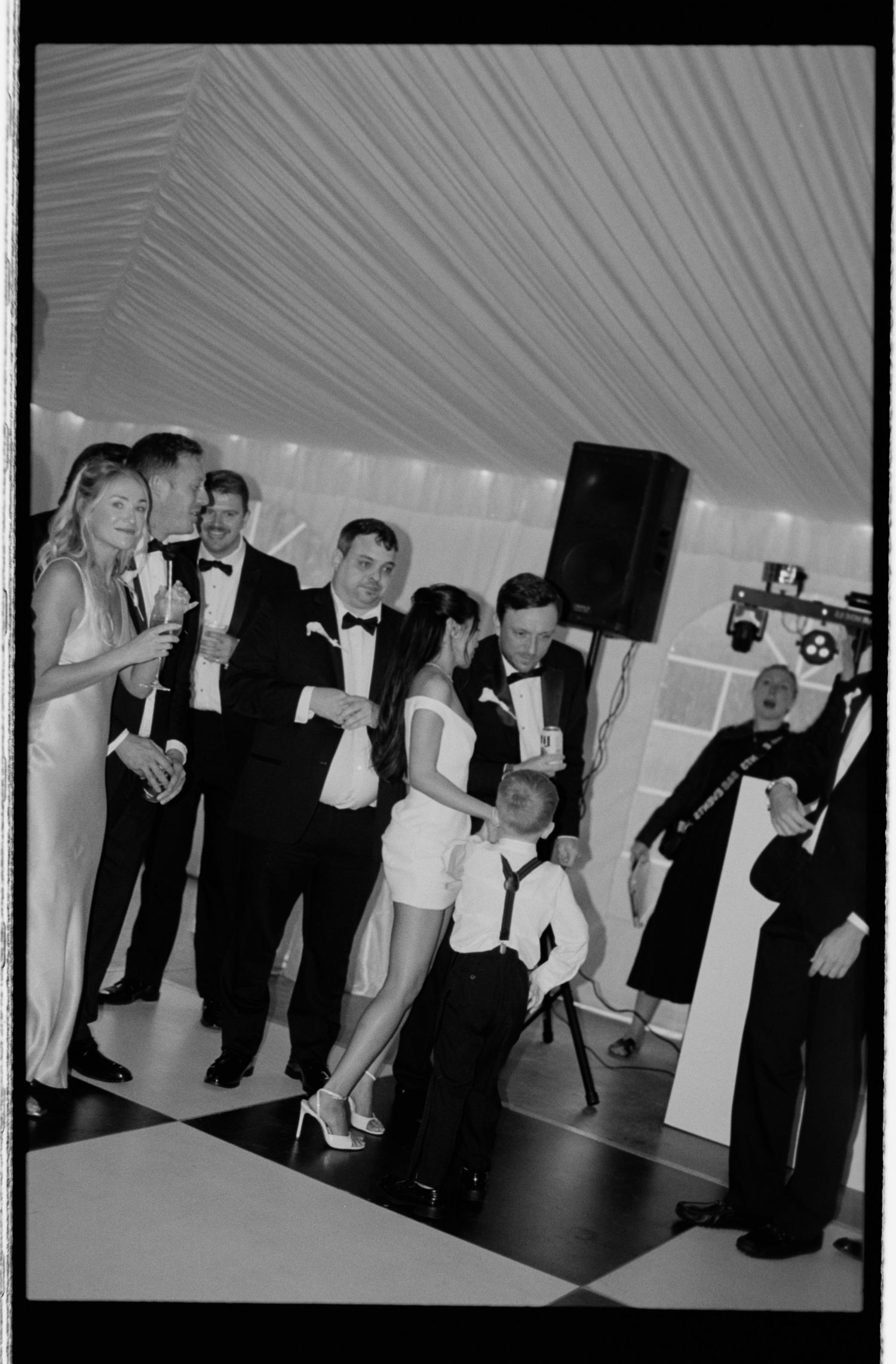 Black and white photo of elegantly dressed wedding guests dancing and socializing on a checkered dance floor at a formal reception.