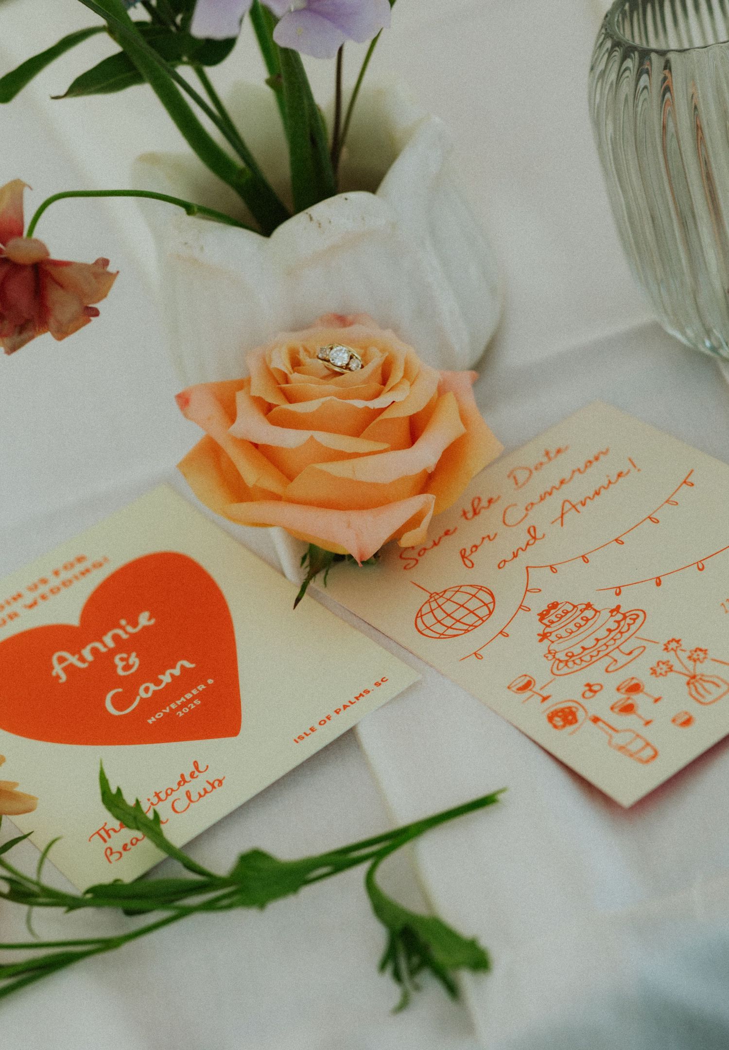 Wedding save-the-date cards for Annie and Cam beside a peach rose with a diamond ring on a white tablecloth.