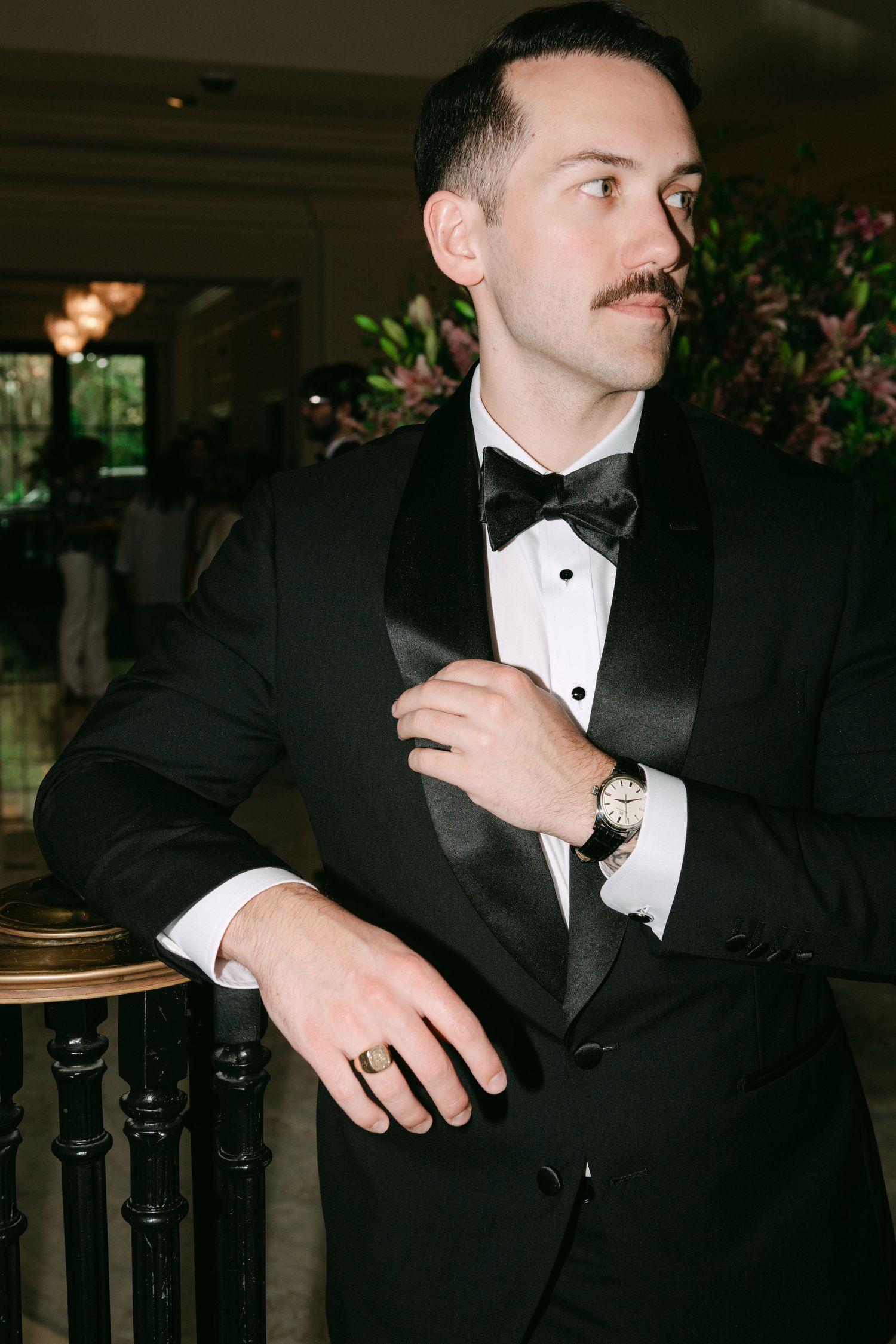 Groom in black tuxedo with bow tie and mustache adjusting jacket at elegant wedding venue.