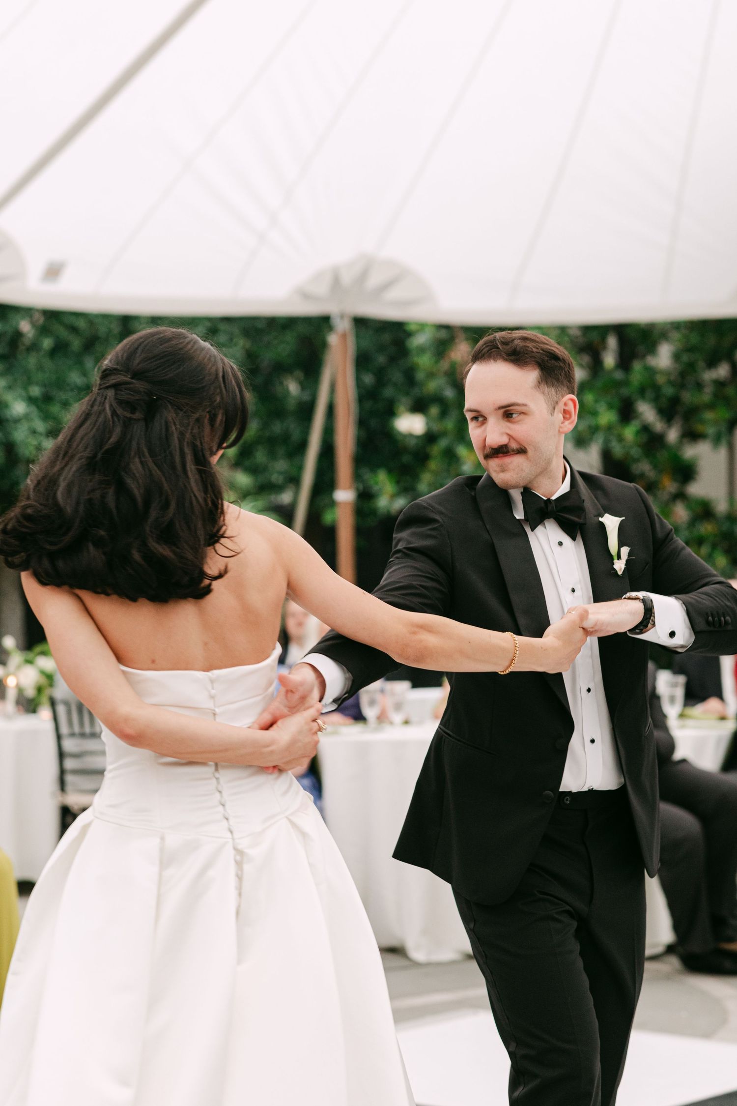 Groom in black tuxedo smiles while dancing with bride in strapless white gown under outdoor tent reception.
