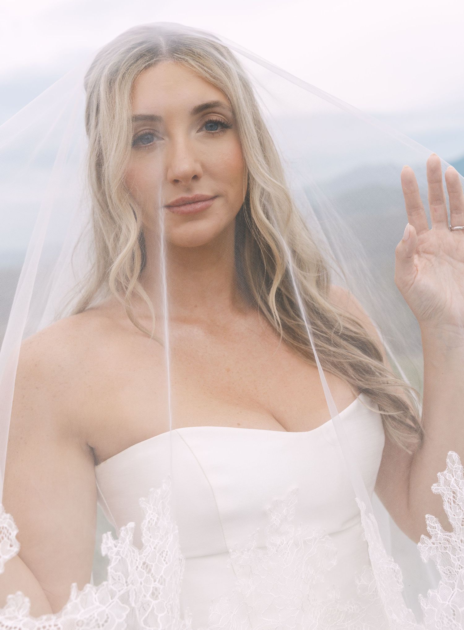 Blonde bride in strapless white gown and sheer veil poses outdoors with mountain views behind her.