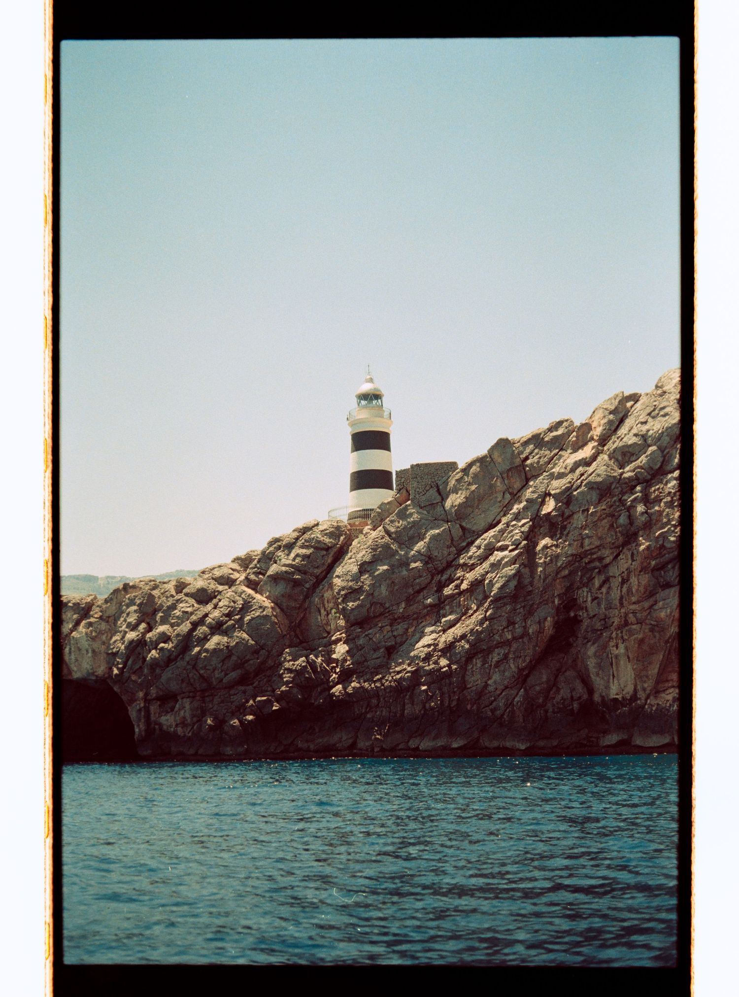 Black and white striped lighthouse perched on rocky coastal cliffs above blue sea waters under a clear sky.