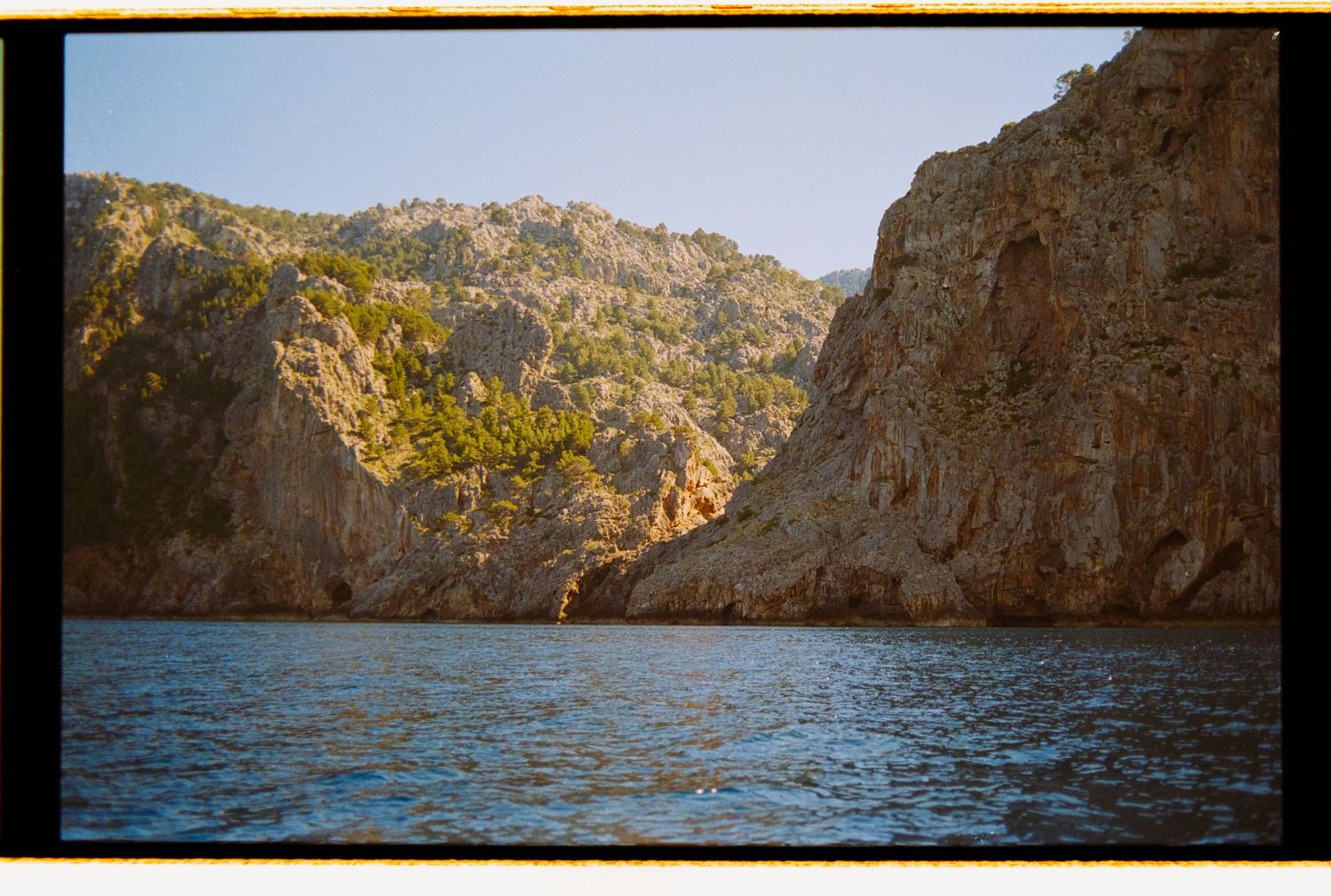 Rocky coastal cliffs with lush green trees frame a calm blue sea inlet under a clear sky.