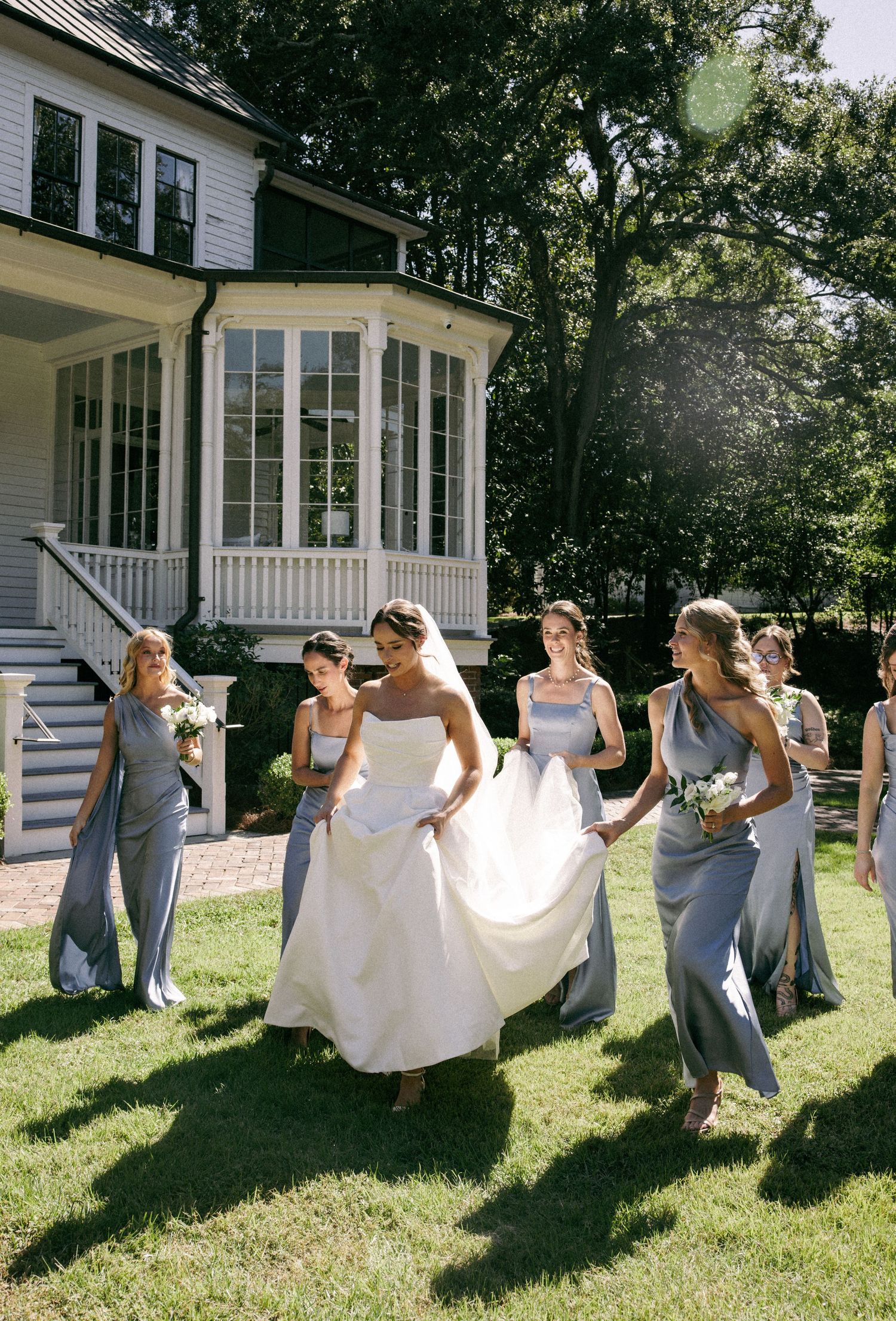 Bride in white gown walks with bridesmaids in silver dresses on green lawn outside elegant white house.