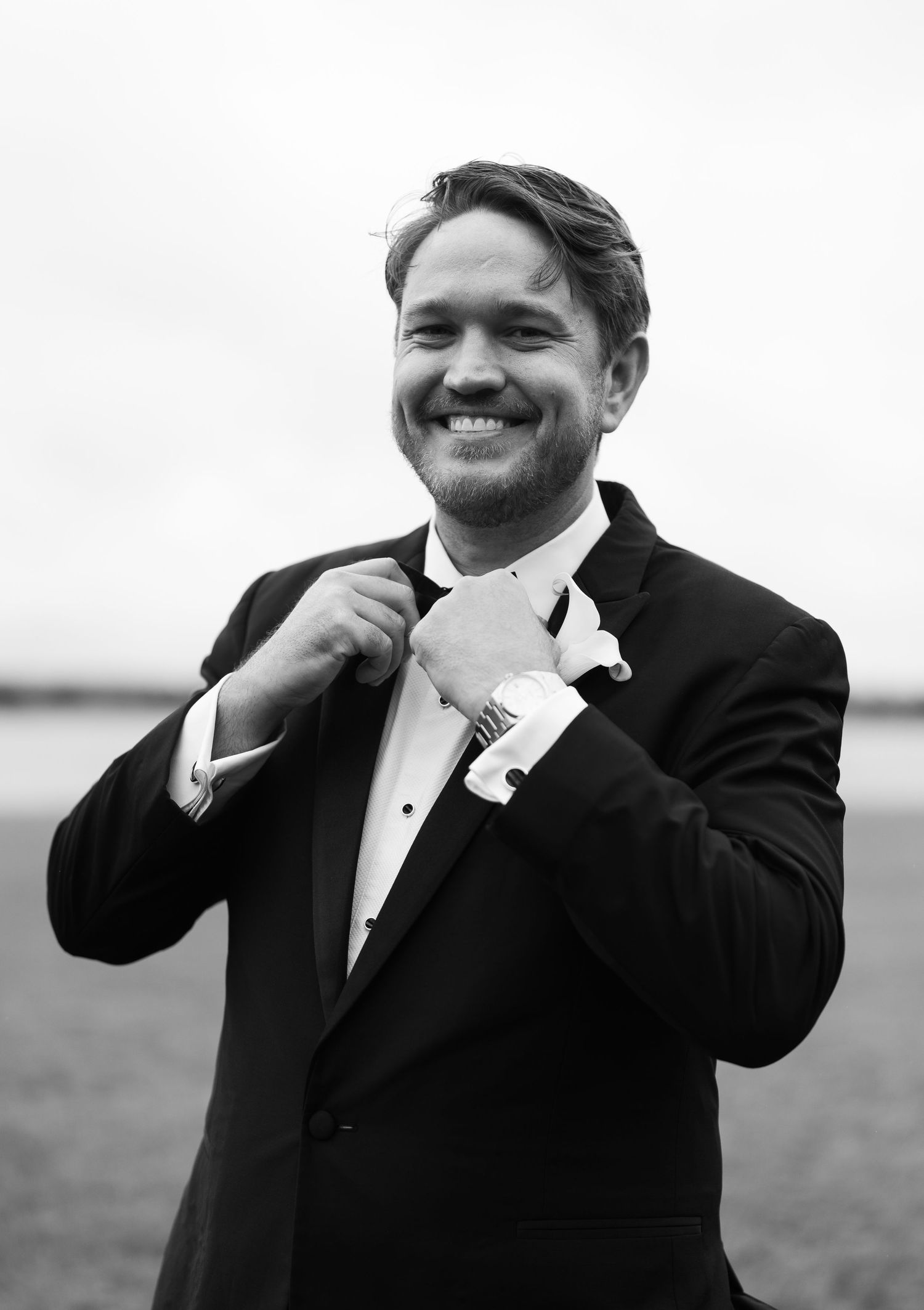 Smiling groom in black tuxedo adjusting bow tie outdoors in black and white wedding portrait.
