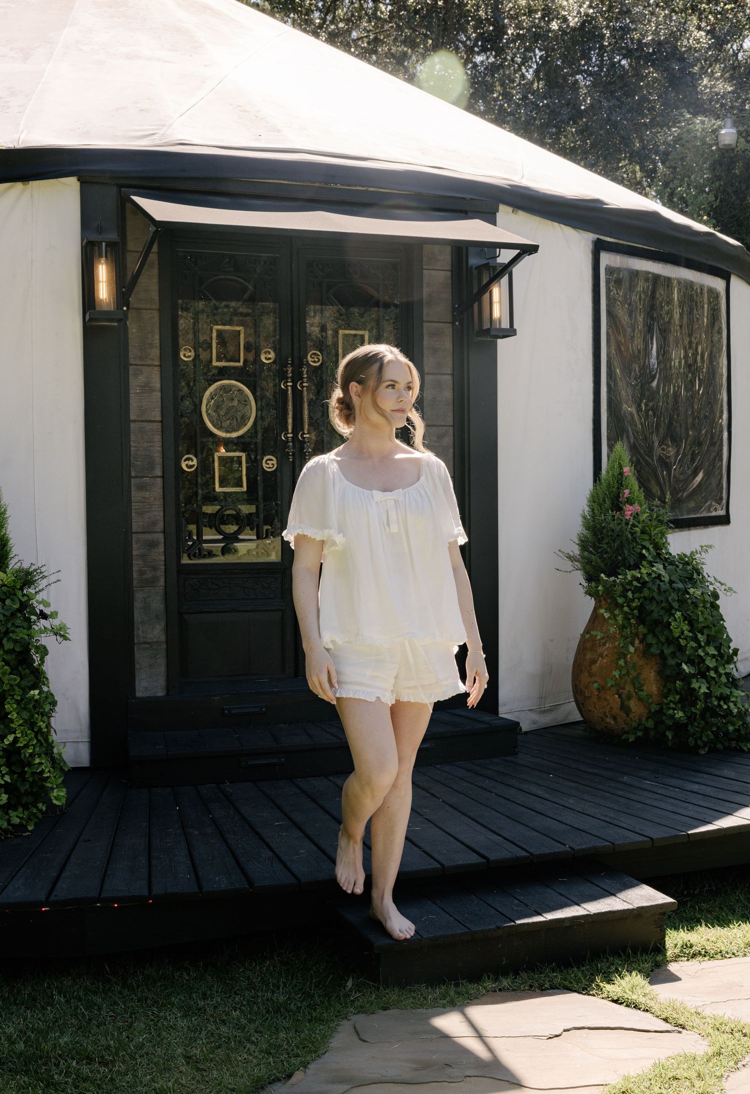 Woman in white top and shorts standing on dark wood deck outside a charming white yurt with black trim.