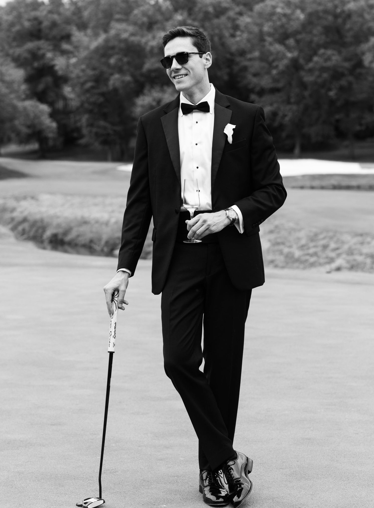 Groom in black tuxedo and bow tie holds golf club on course, smiling in sunglasses with boutonniere.