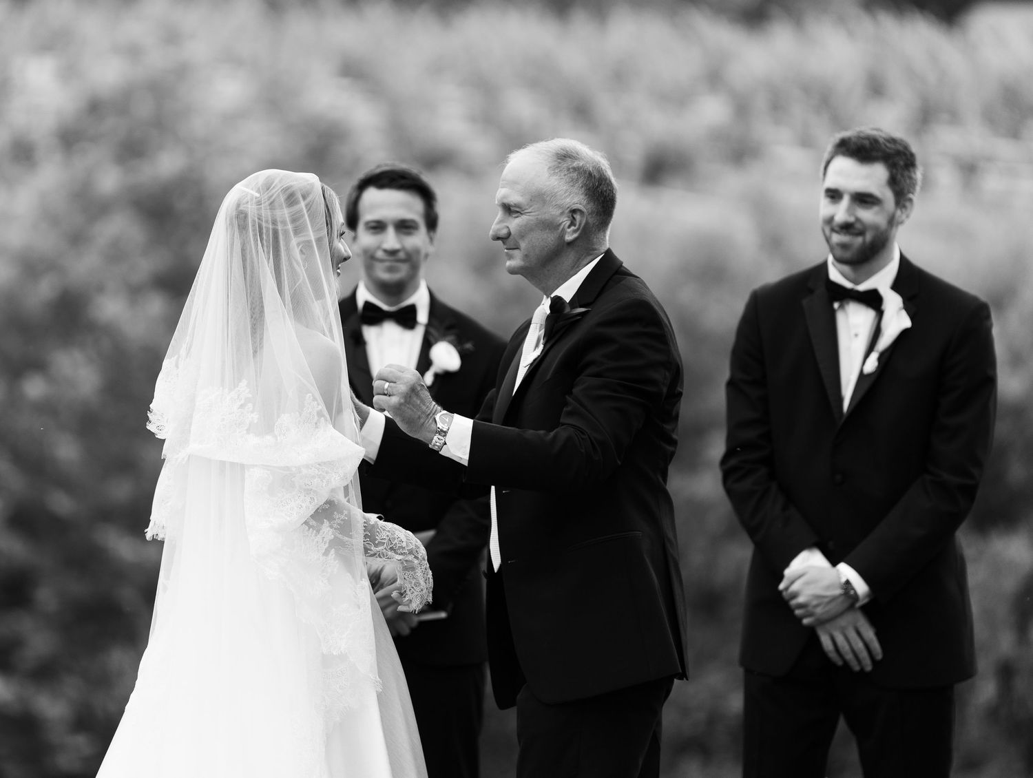 Black and white wedding photo of bride with veiled headpiece sharing a tender moment with older gentleman in tuxedo.