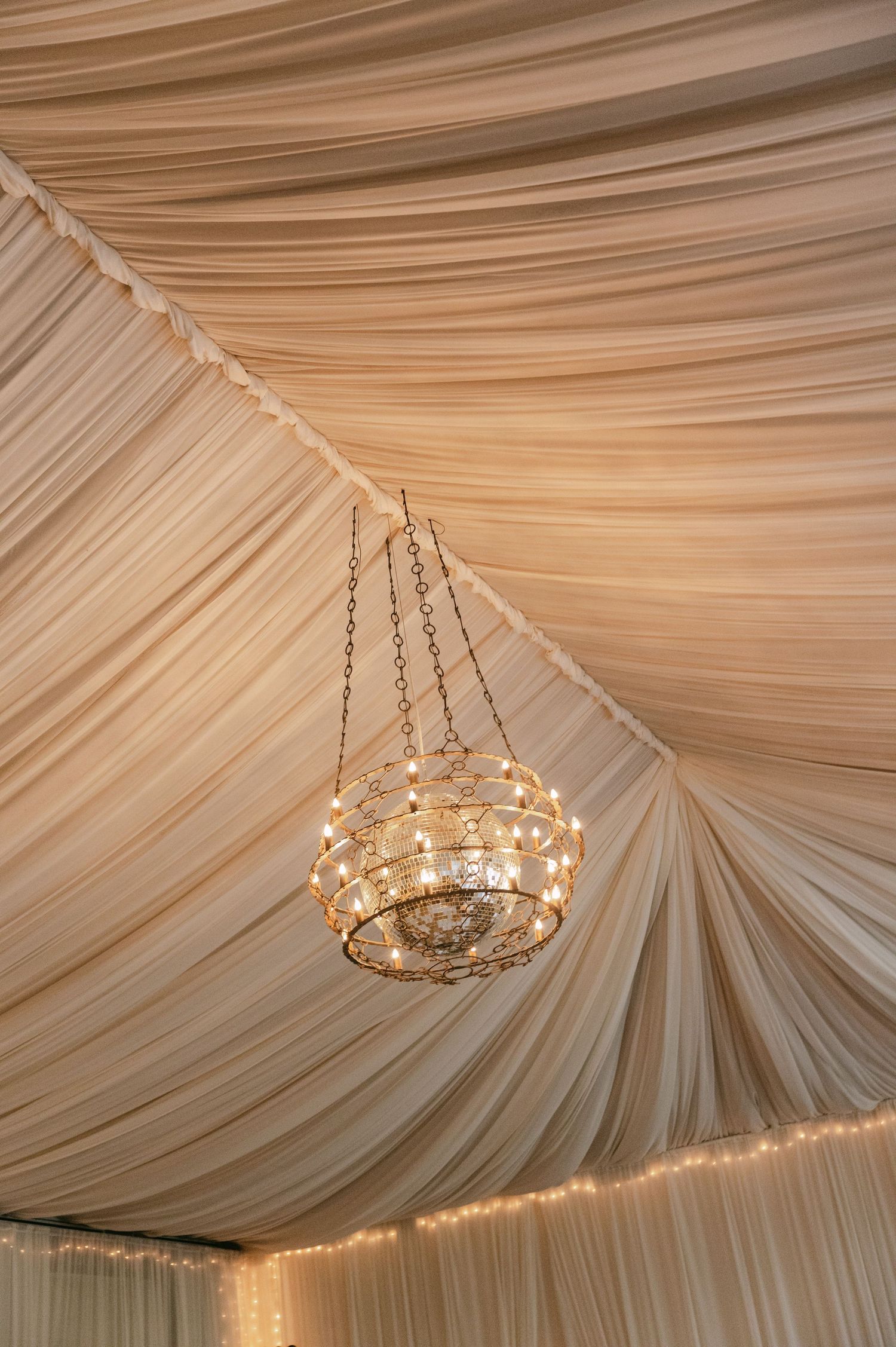 Elegant crystal chandelier hanging from draped ivory fabric ceiling with string lights at wedding venue.