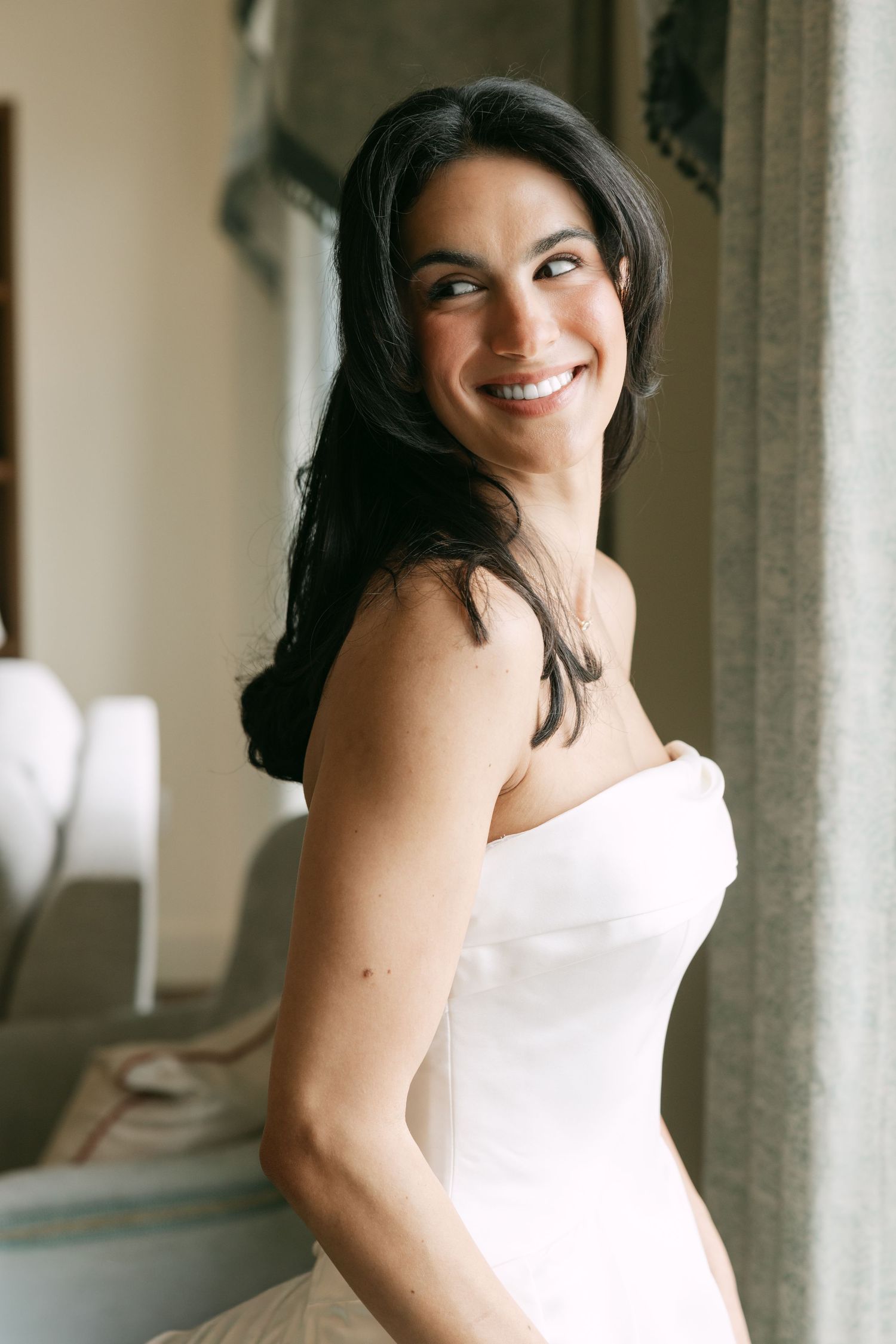 Smiling bride in strapless white wedding gown with dark hair, posing elegantly near a window in a softly lit room.