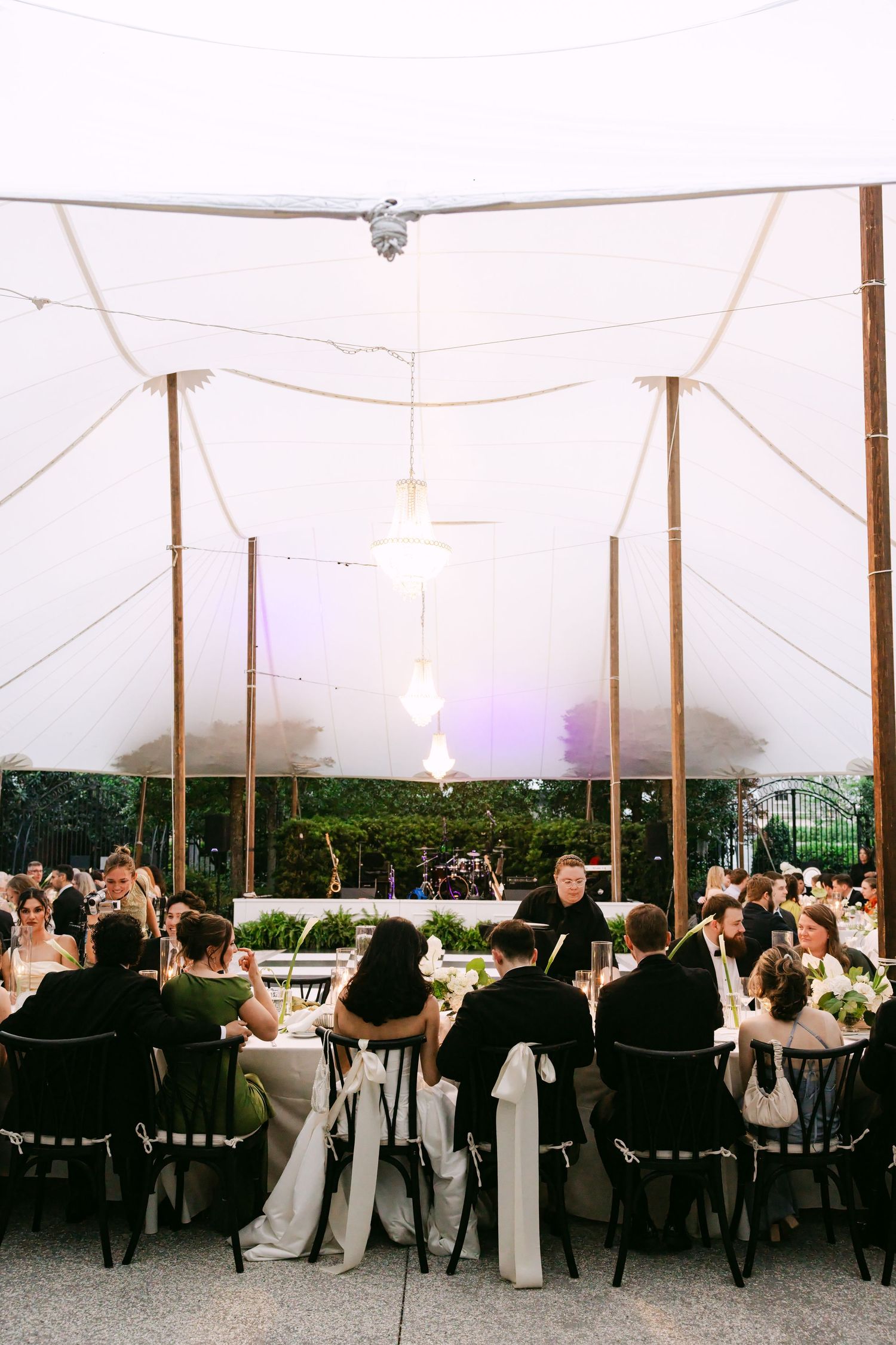 Wedding guests seated at long reception tables under a white sailcloth tent with hanging Edison bulb lights.
