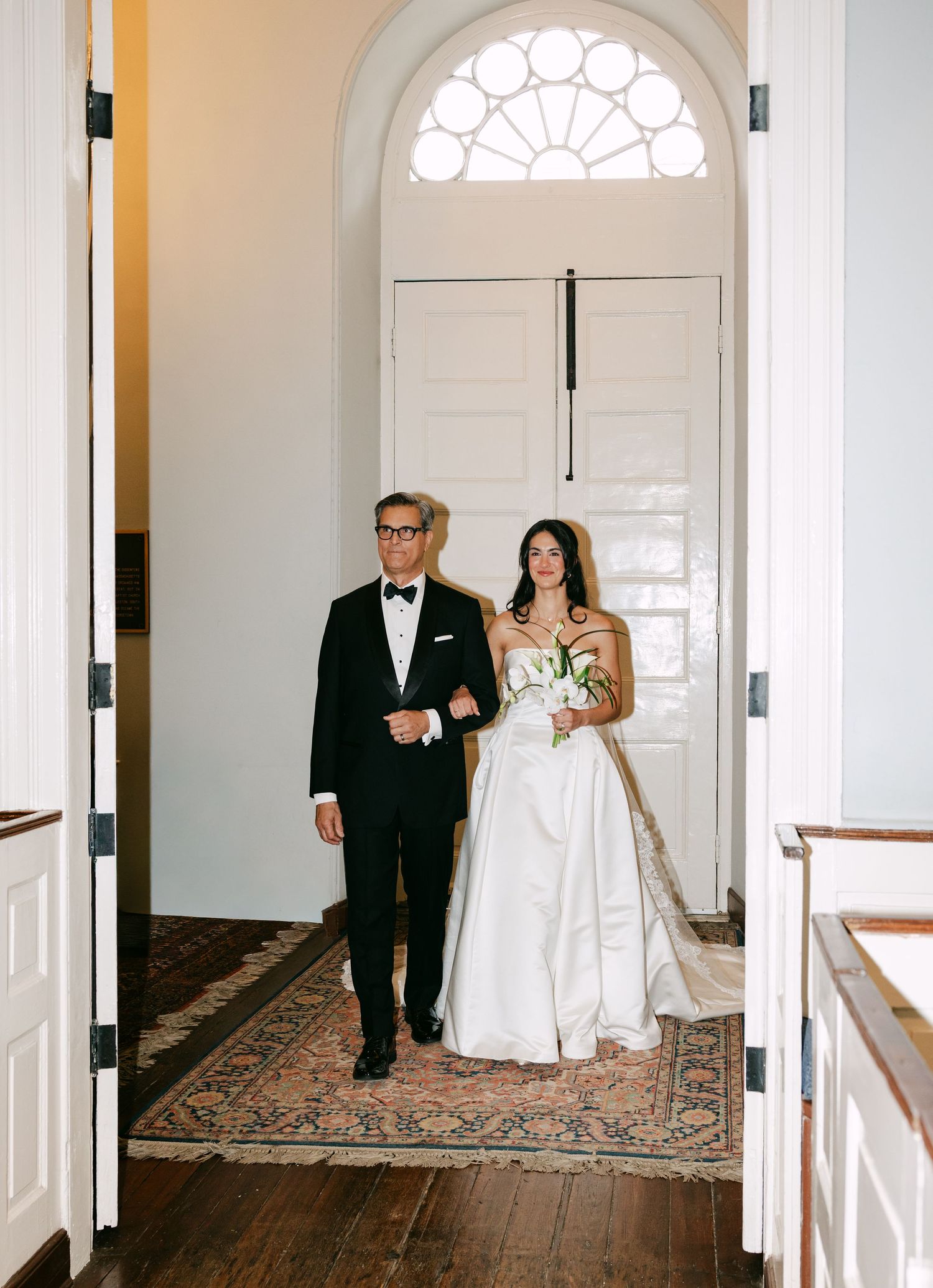 Bride in white gown walks down aisle with escort in tuxedo through elegant white arched doorway.