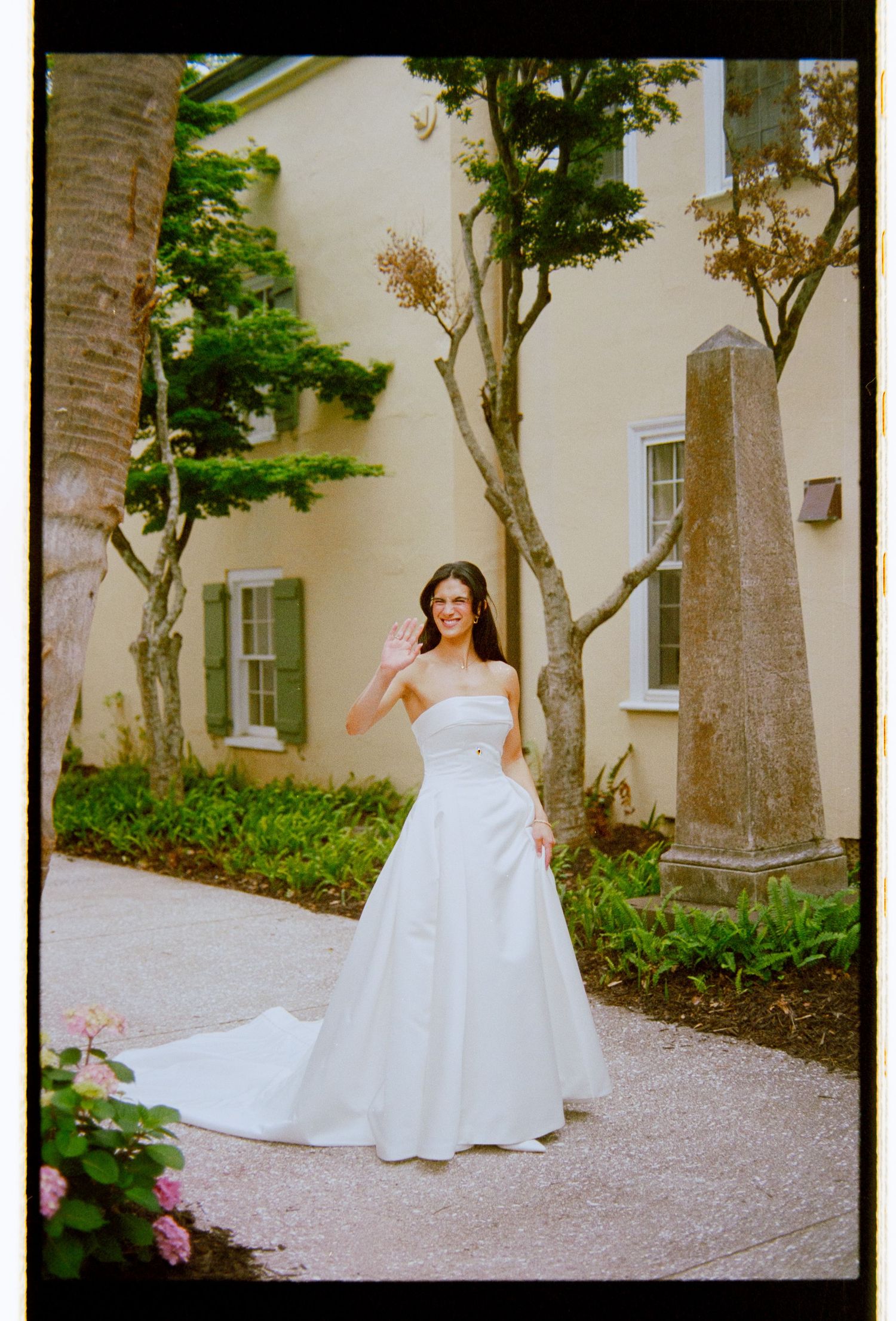 Bride in a strapless white ball gown waves outdoors near a yellow building on her wedding day.